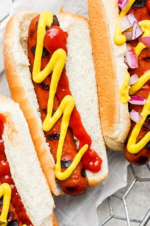 Three smoked hot dogs in buns with various toppings sitting in a wire basket.
