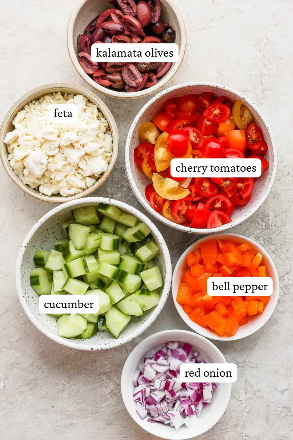 Greek tomato and cucumber salad ingredients in individual bowls of kalamata olives, feta cheese, cherry tomatoes, cucumber, bell pepper, and red onion.