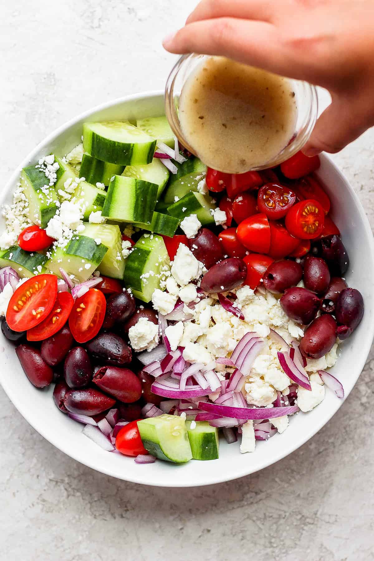 Cucumbers, bell peppers, olives, tomatoes, red onion, and feta cheese in a bowl with Greek vinaigrette being poured on top.