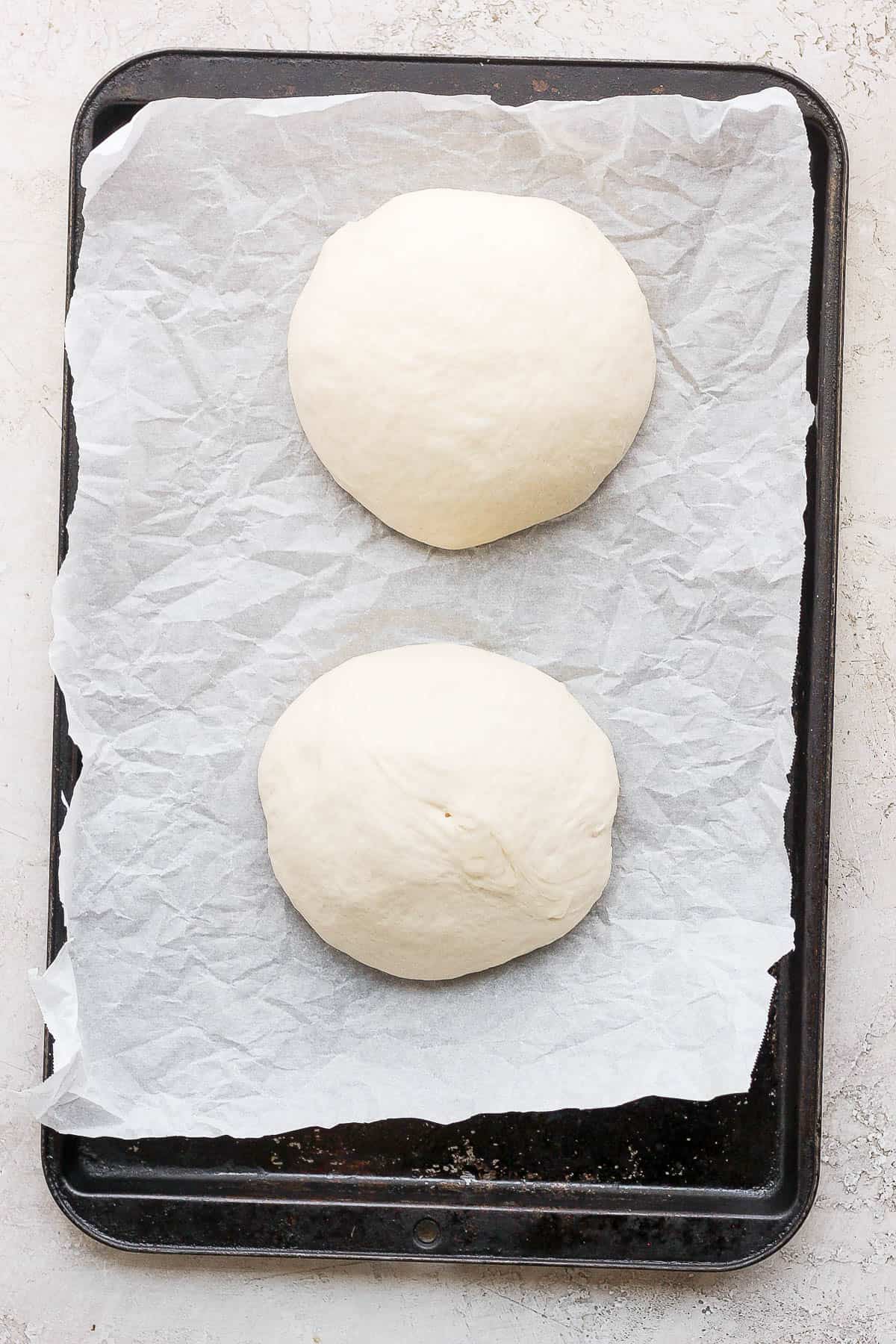 Two portions of pizza dough on parchment-lined sheet. 