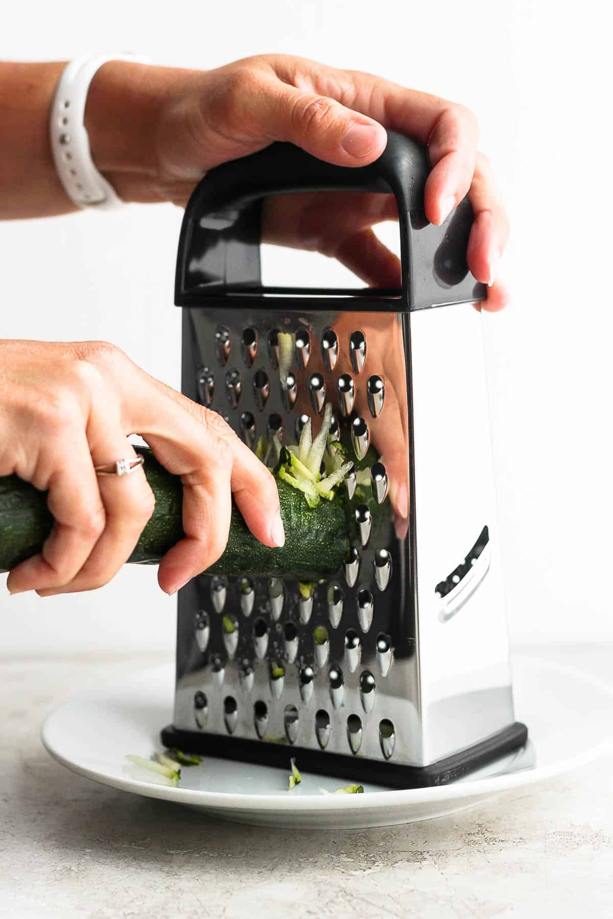 Zucchini being grated on a box grater over a white plate.