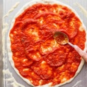 Rolled out pizza dough on a pan and someone is spreading pizza sauce all over it with the back of a spoon.