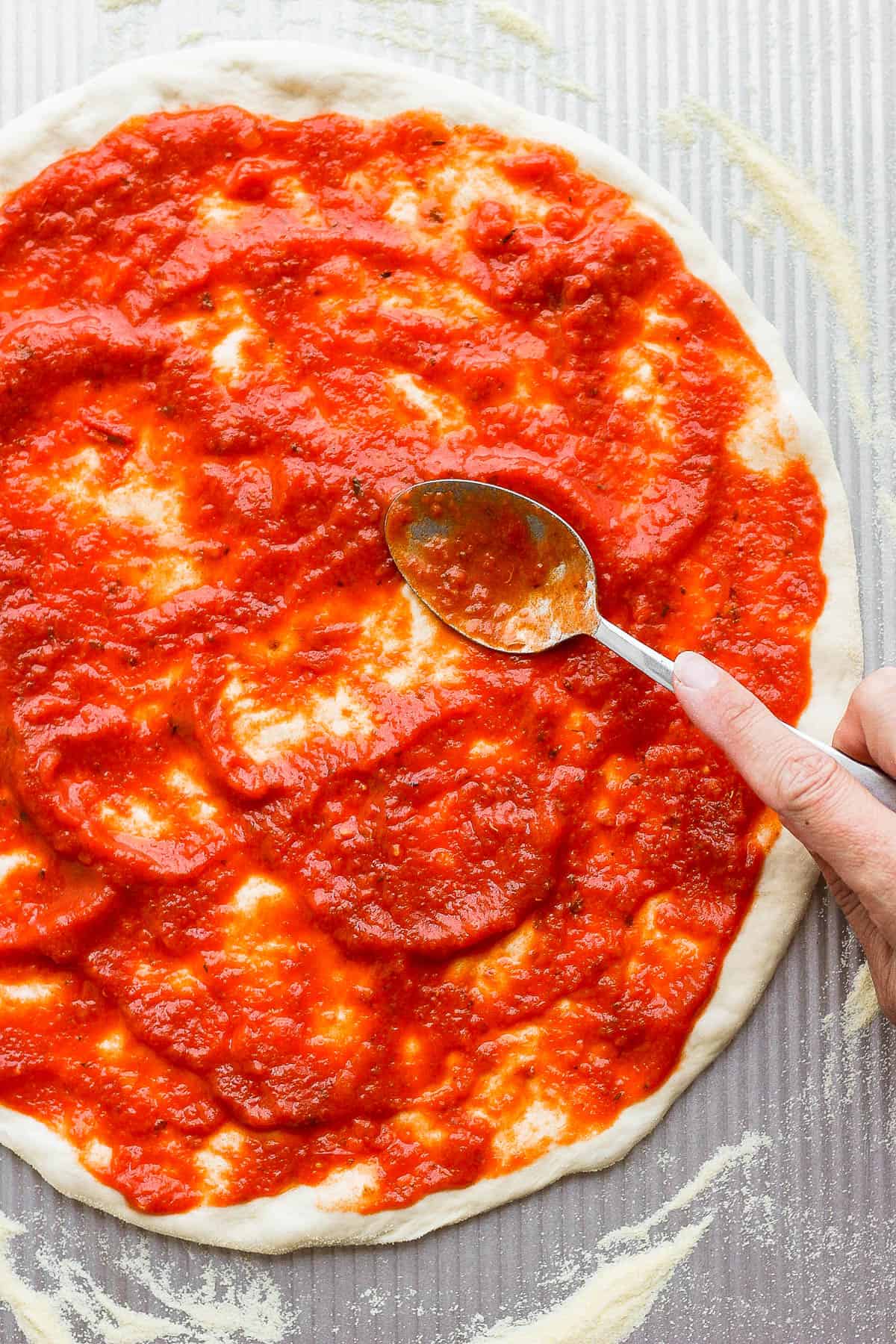 Pizza sauce being spread on homemade dough with a spoon.