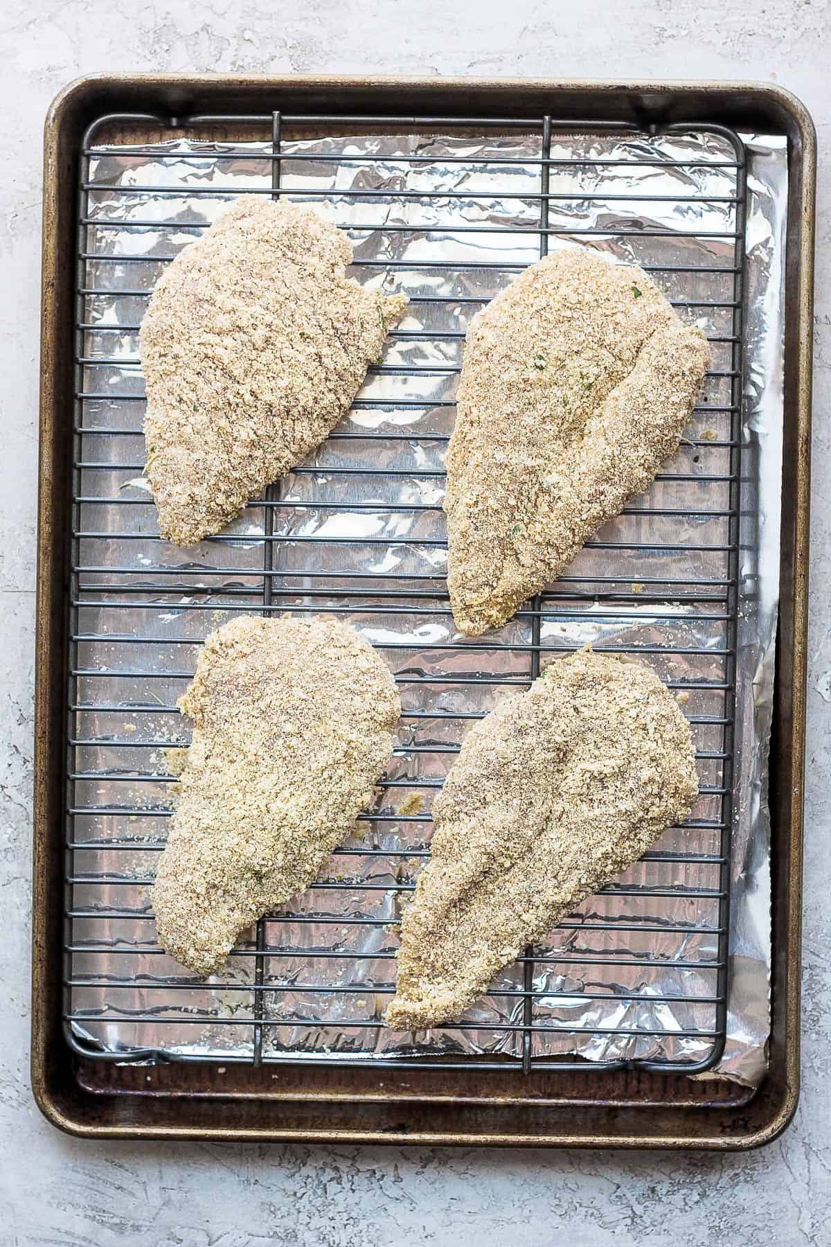 A baking sheet with a piece of tinfoil on the bottom, topped with a baking rack with four breaded chicken breasts on top.