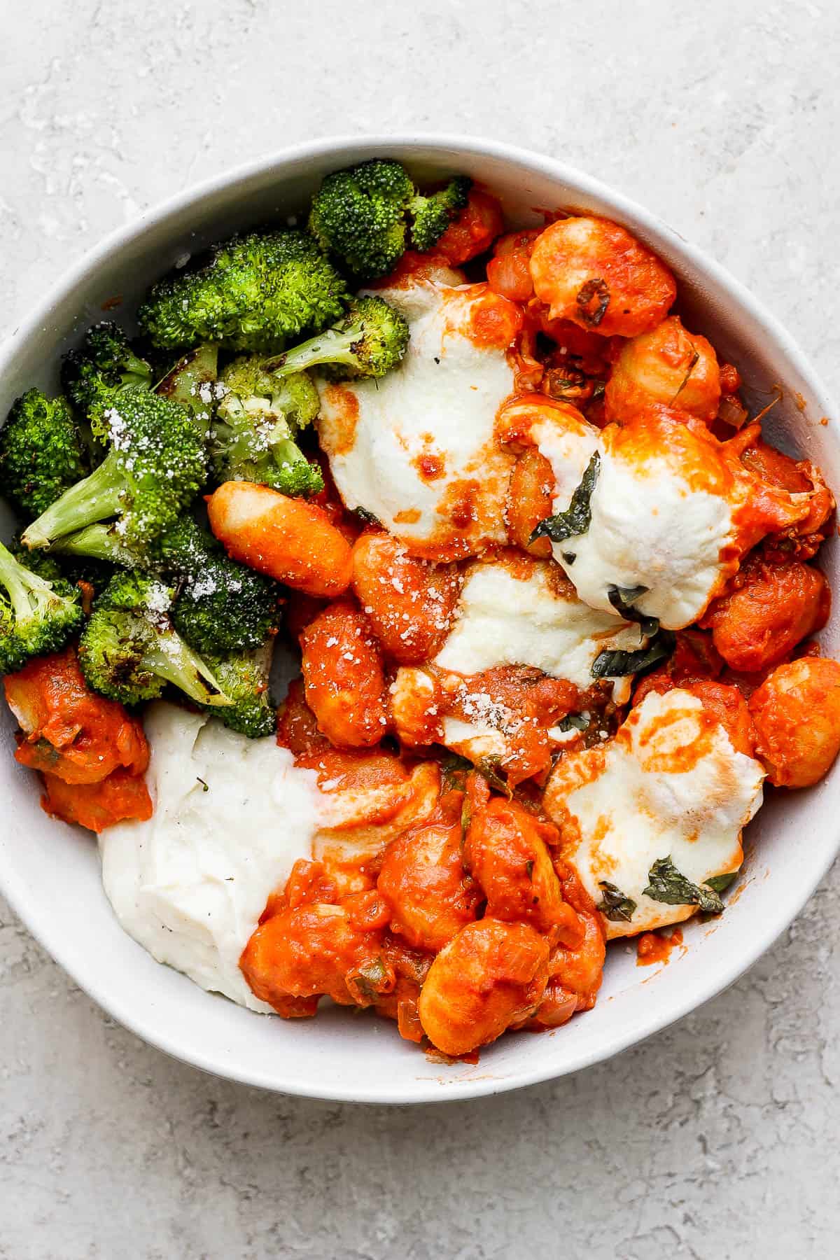 A bowl of baked gnocchi with a side of grilled broccoli.