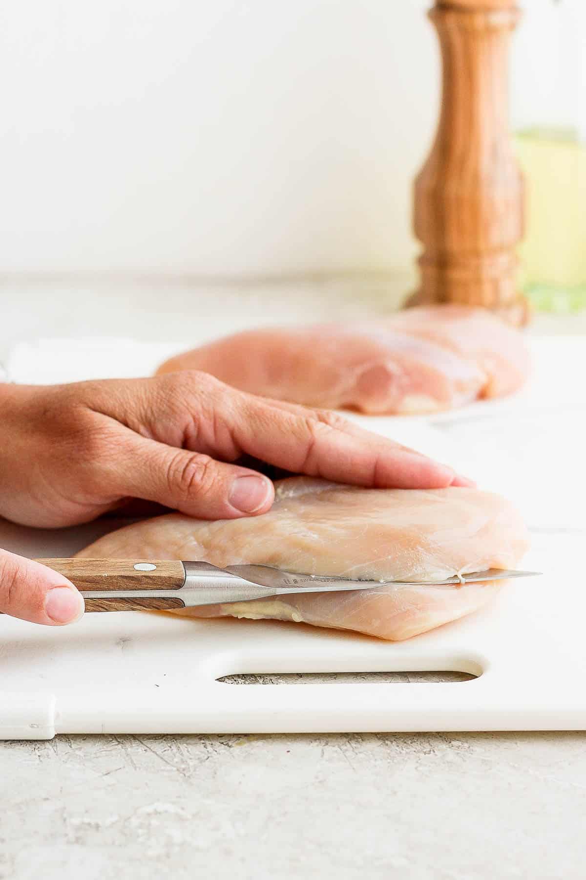 A hand placed on top of a raw chicken breast while the other cuts through the middle of the chicken breast lengthwise to the otherside. 