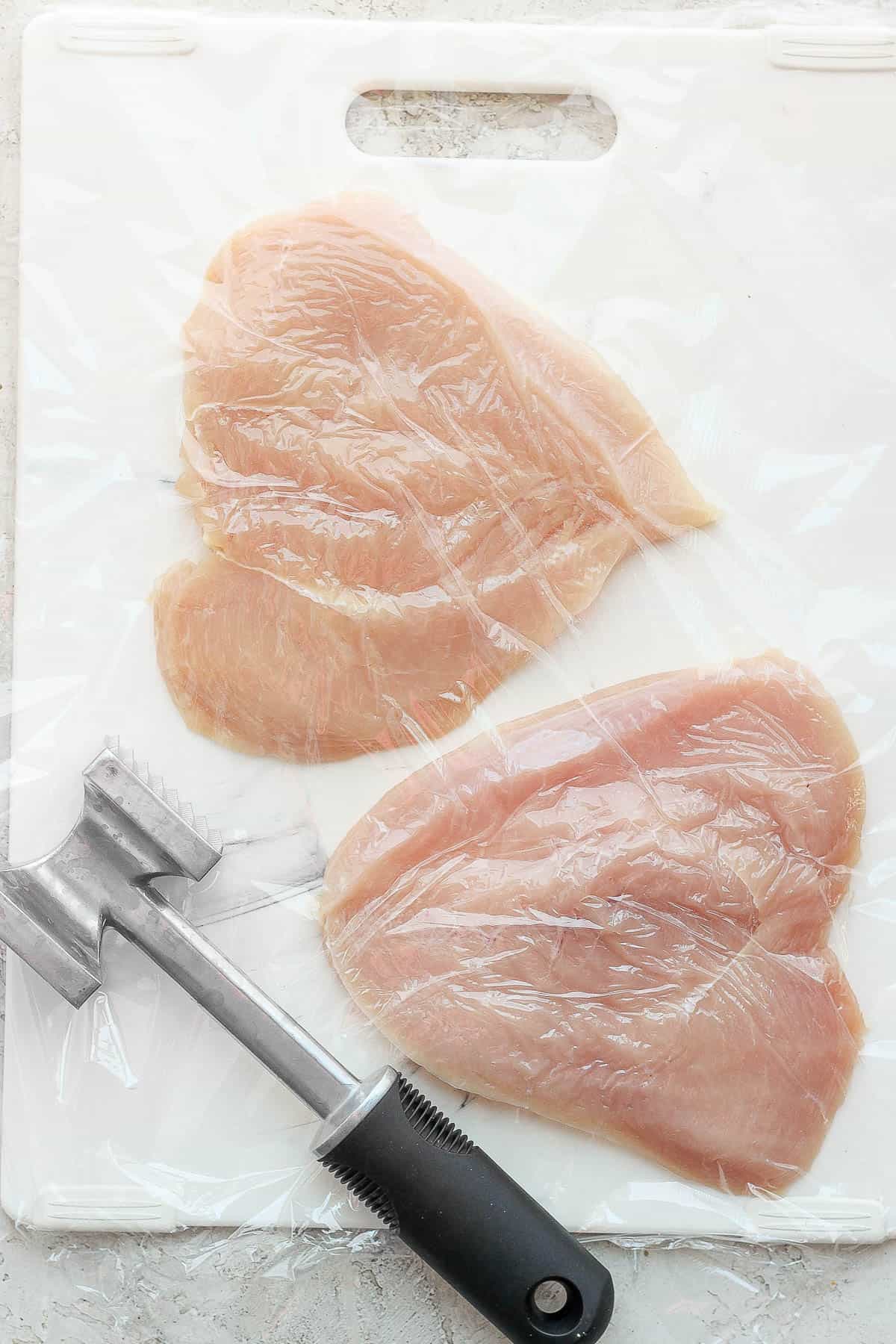 An overhead view of two butterflied chicken breasts on a cutting board with plastic wrap over the top and a meat tenderizer to the side.