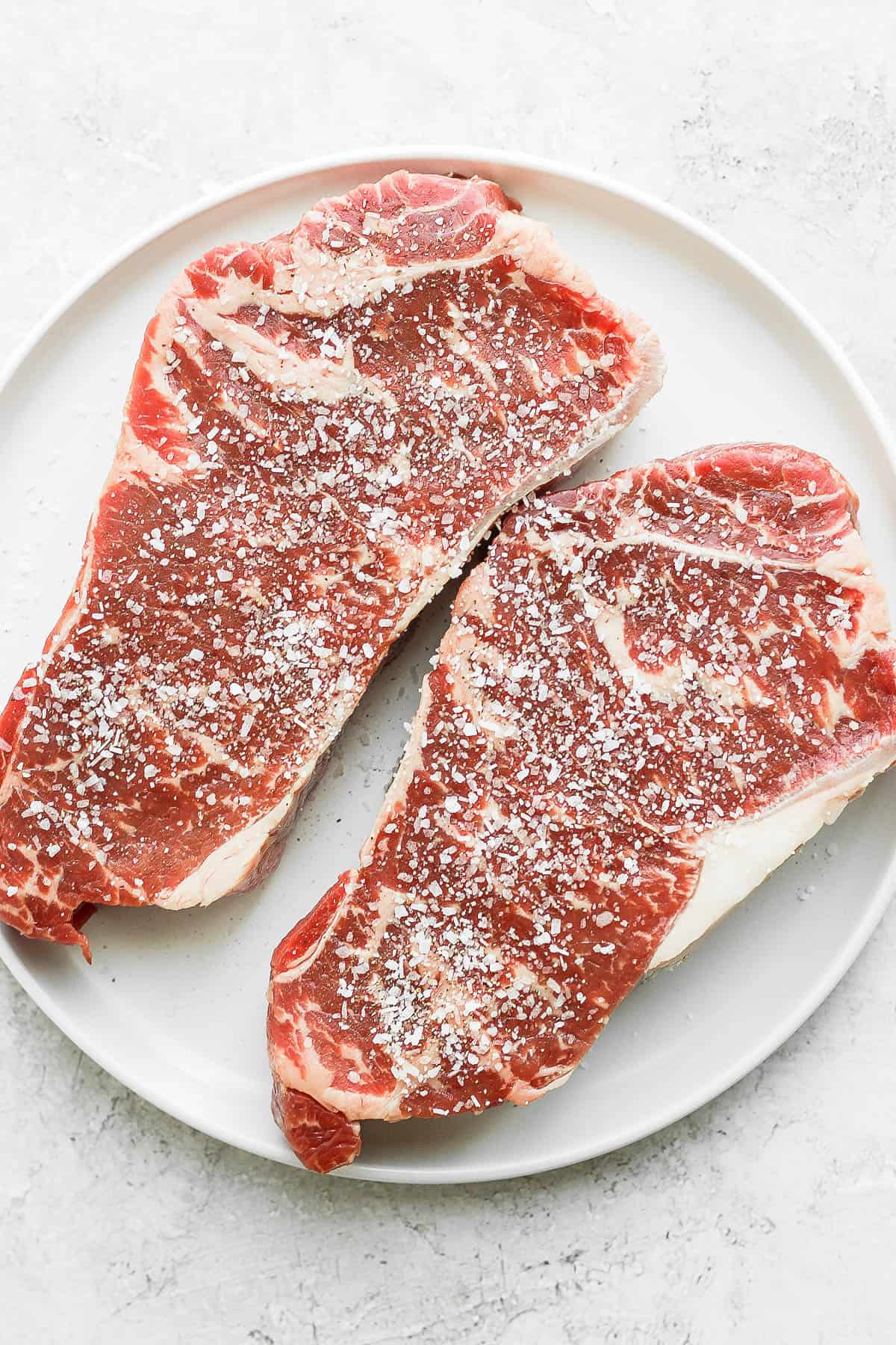 NY strip steaks on a plate seasoned with salt and pepper.