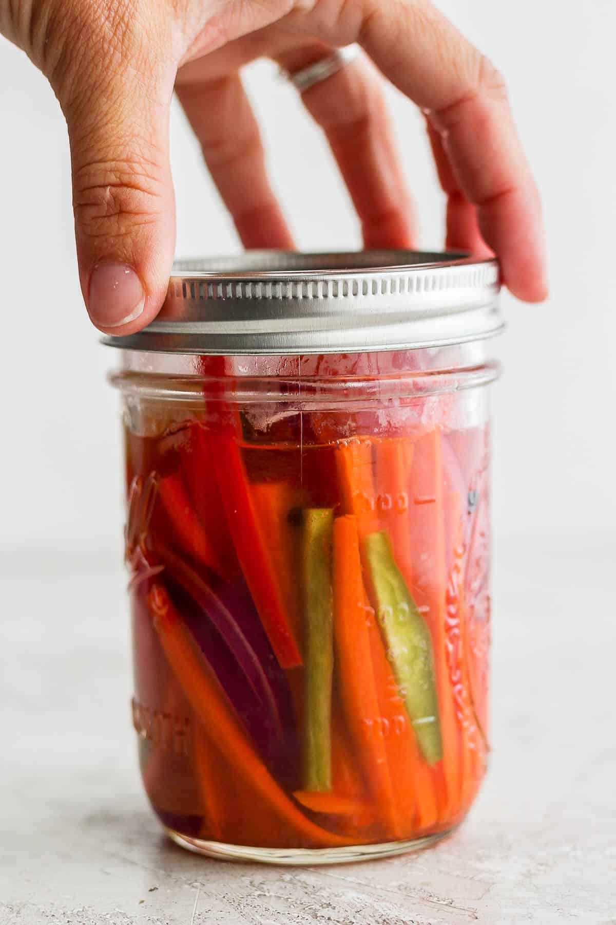 A hand placing the lid on a mason jar of quick pickled vegetables.