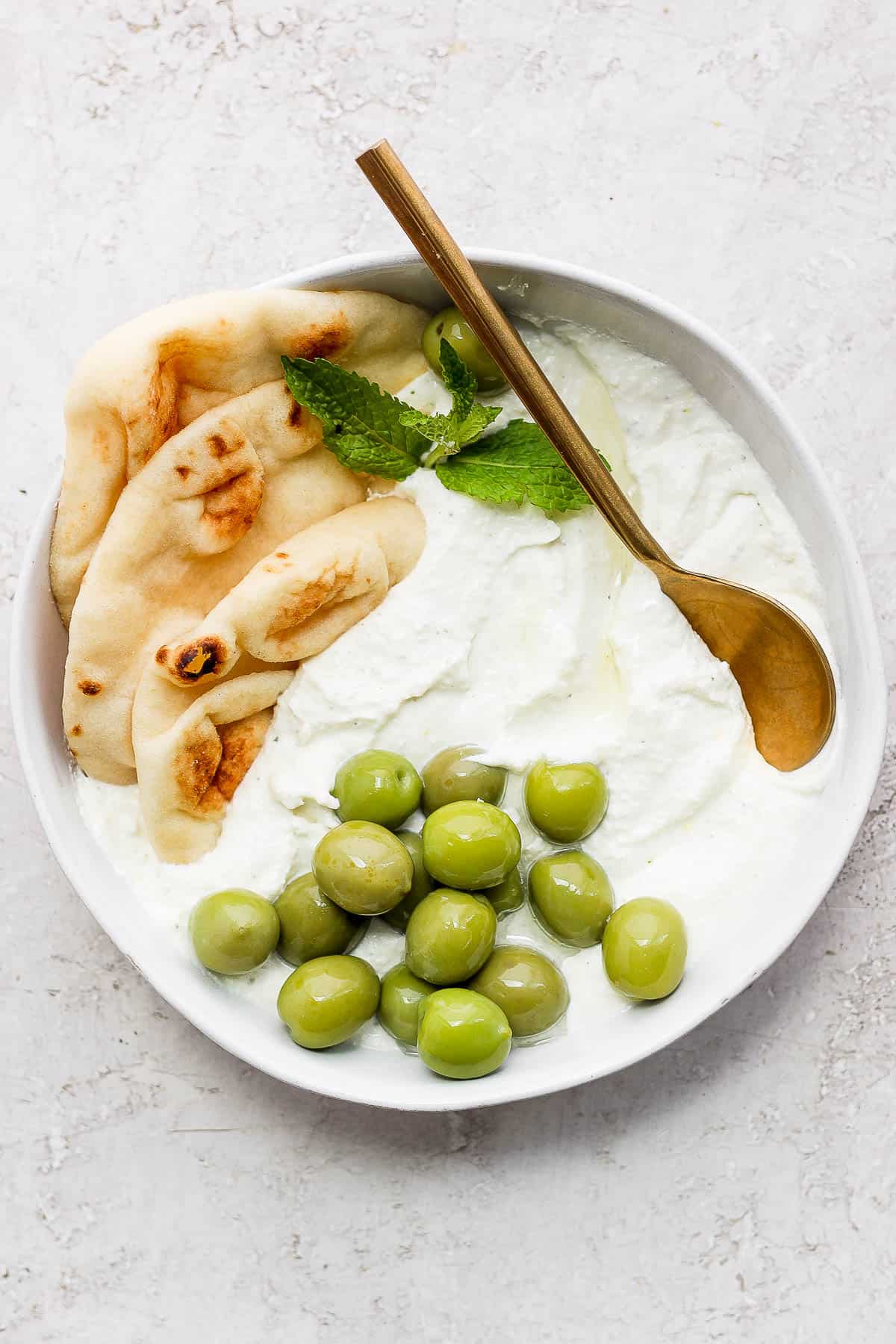 Whipped feta dip in a bowl surrounded by pita bread, olives, and a mint leaf.