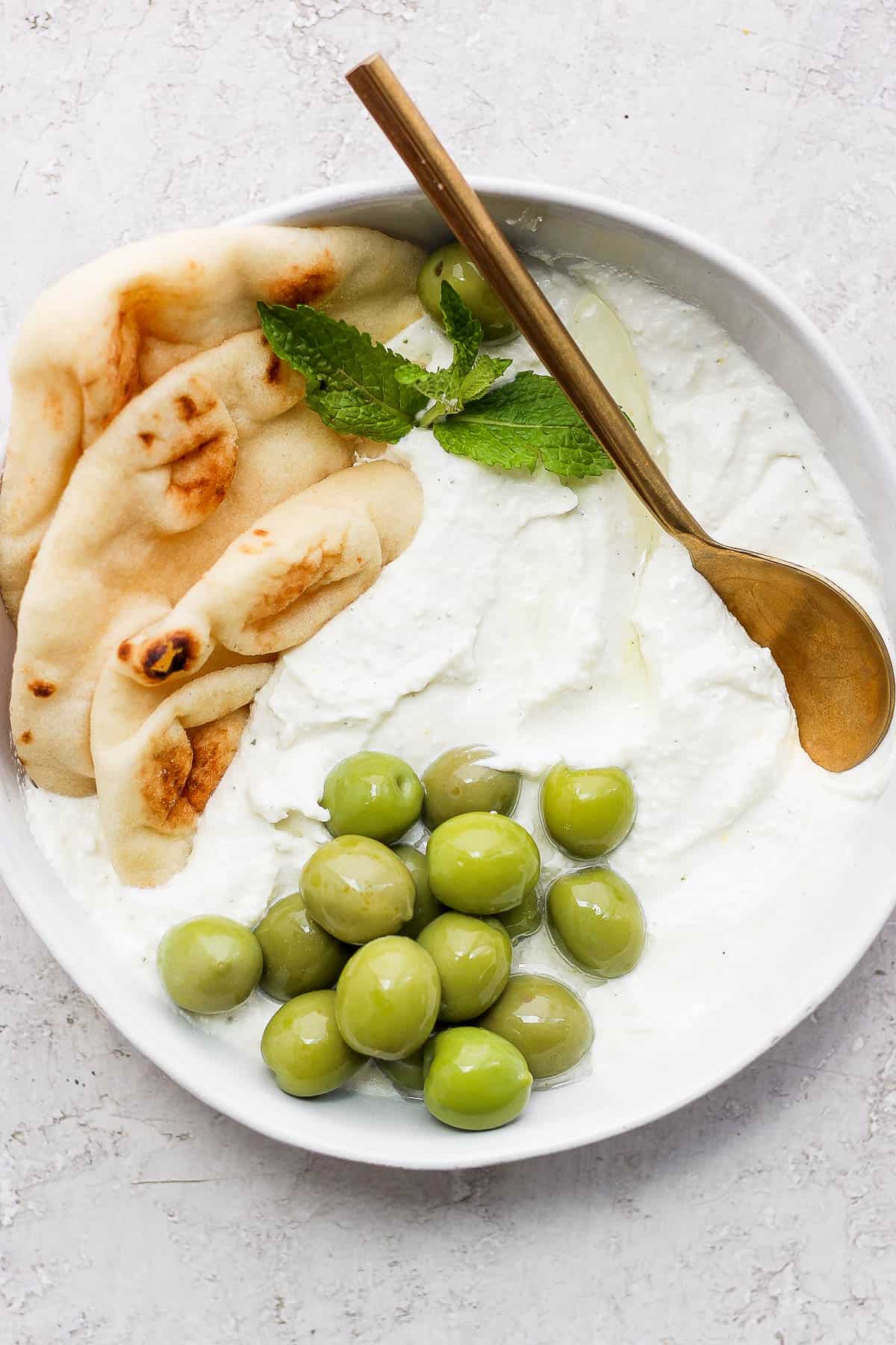 Whipped feta in a bowl accompanied by three pieces of pita bread, green olives, and a few mint leaves.