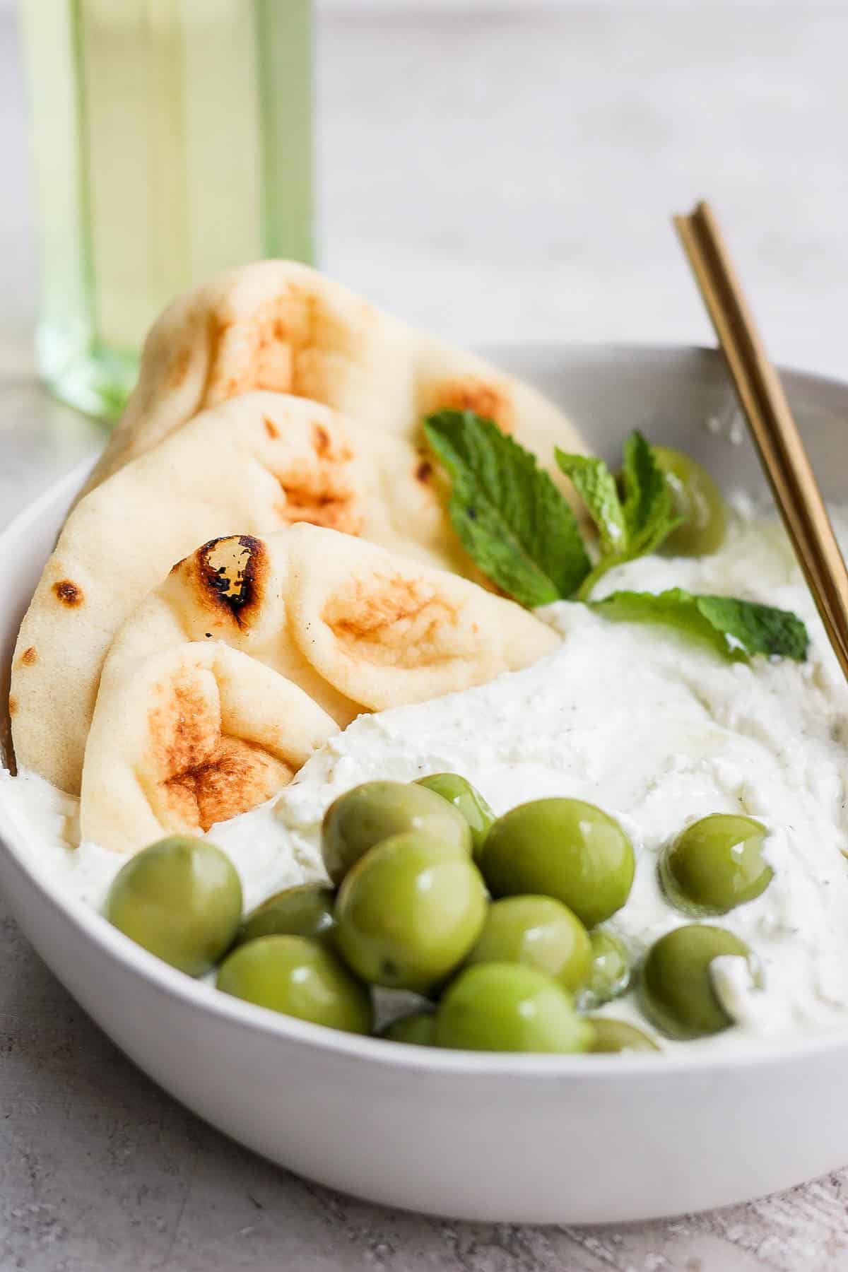 A close up of the whipped feta in a bowl accompanied by pita bread, green olives, and mint. 