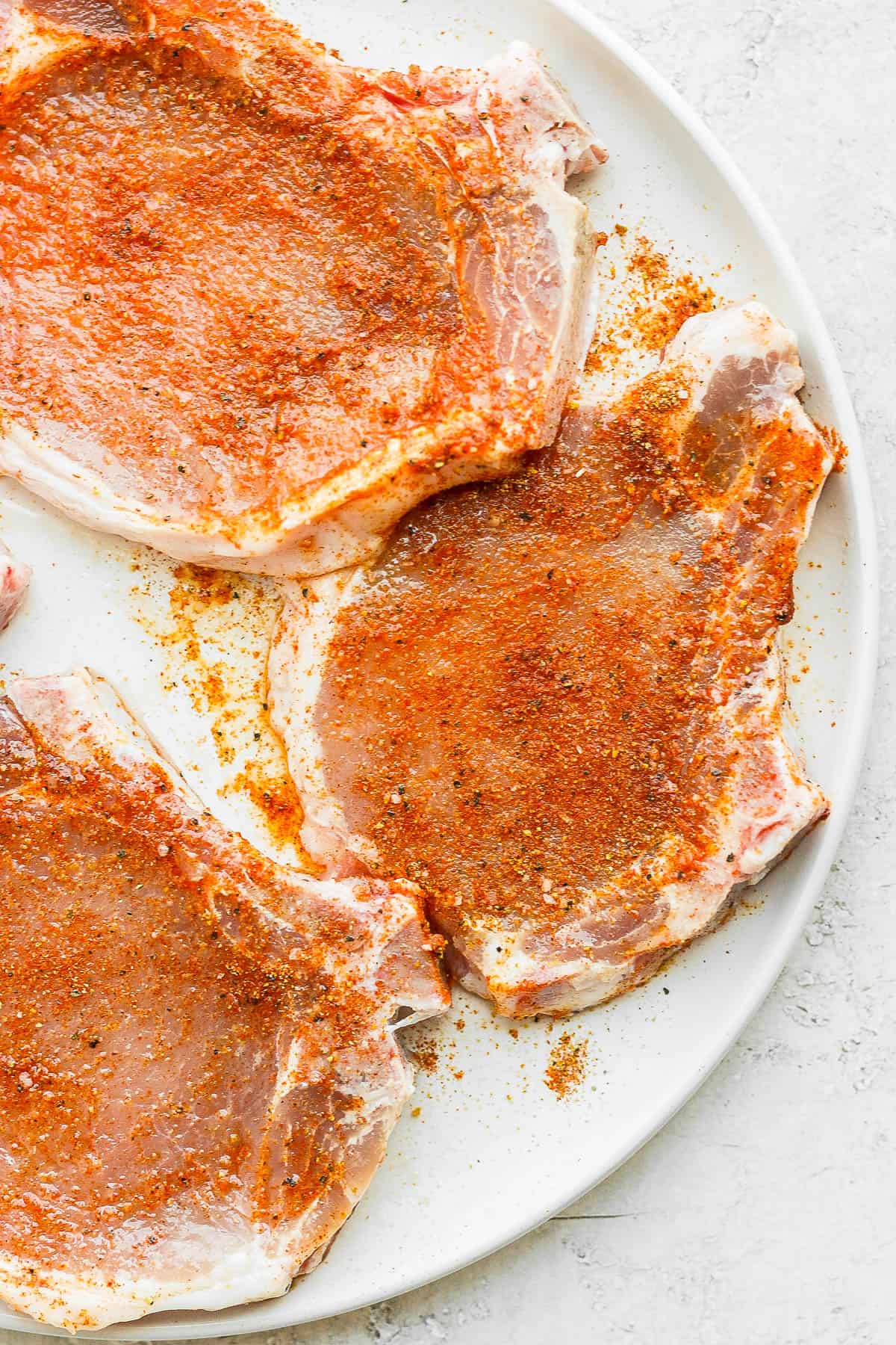Pork chops covered with olive oil and seasoning mixture on a white plate. 