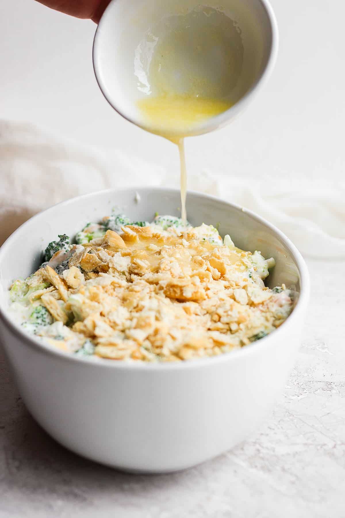 Melted butter being poured out of a small bowl onto the crushed Ritz crackers topping the casserole.