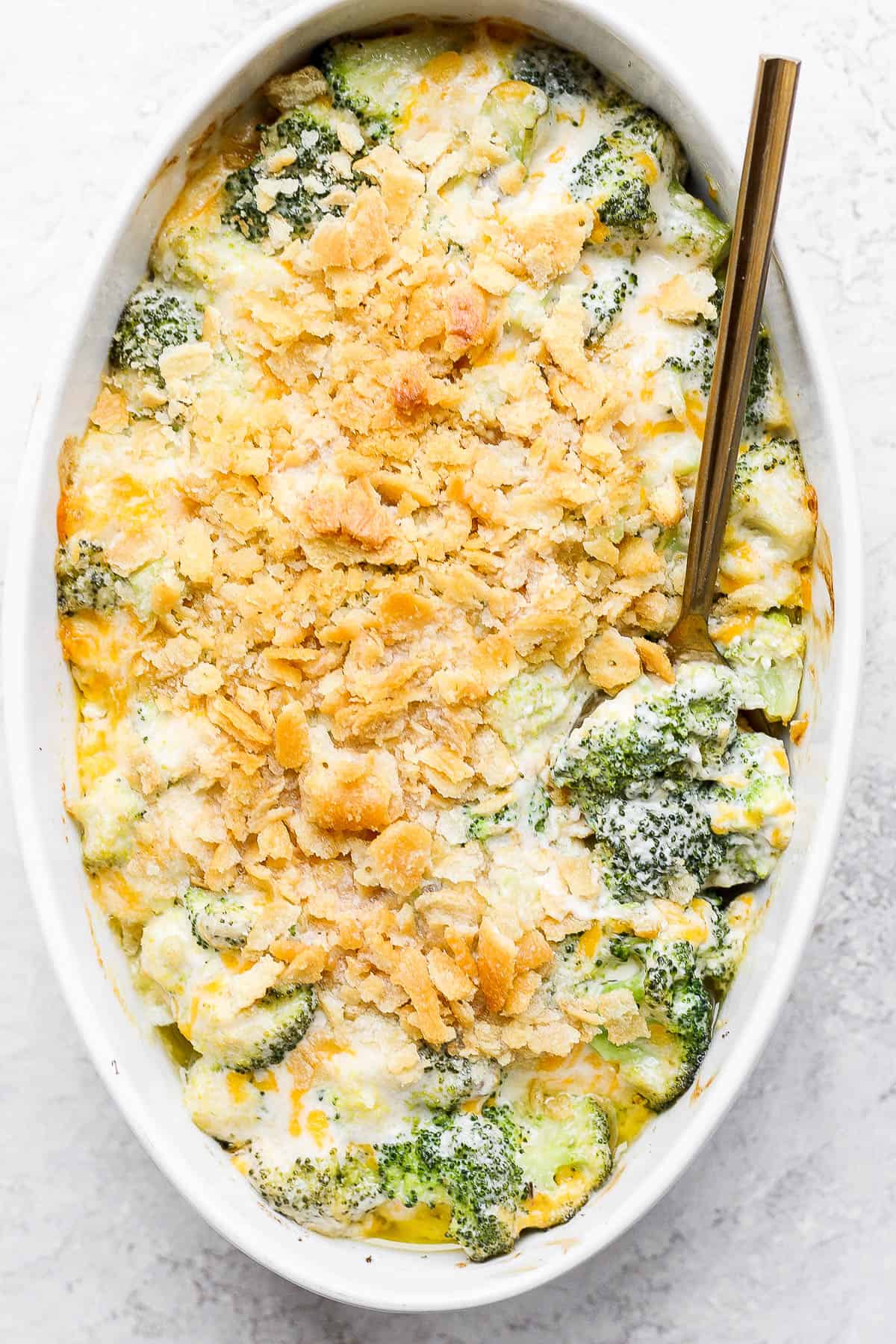 An oval casserole dish filled with baked broccoli casserole with a spoon sticking into it and leaning on the side of the dish.