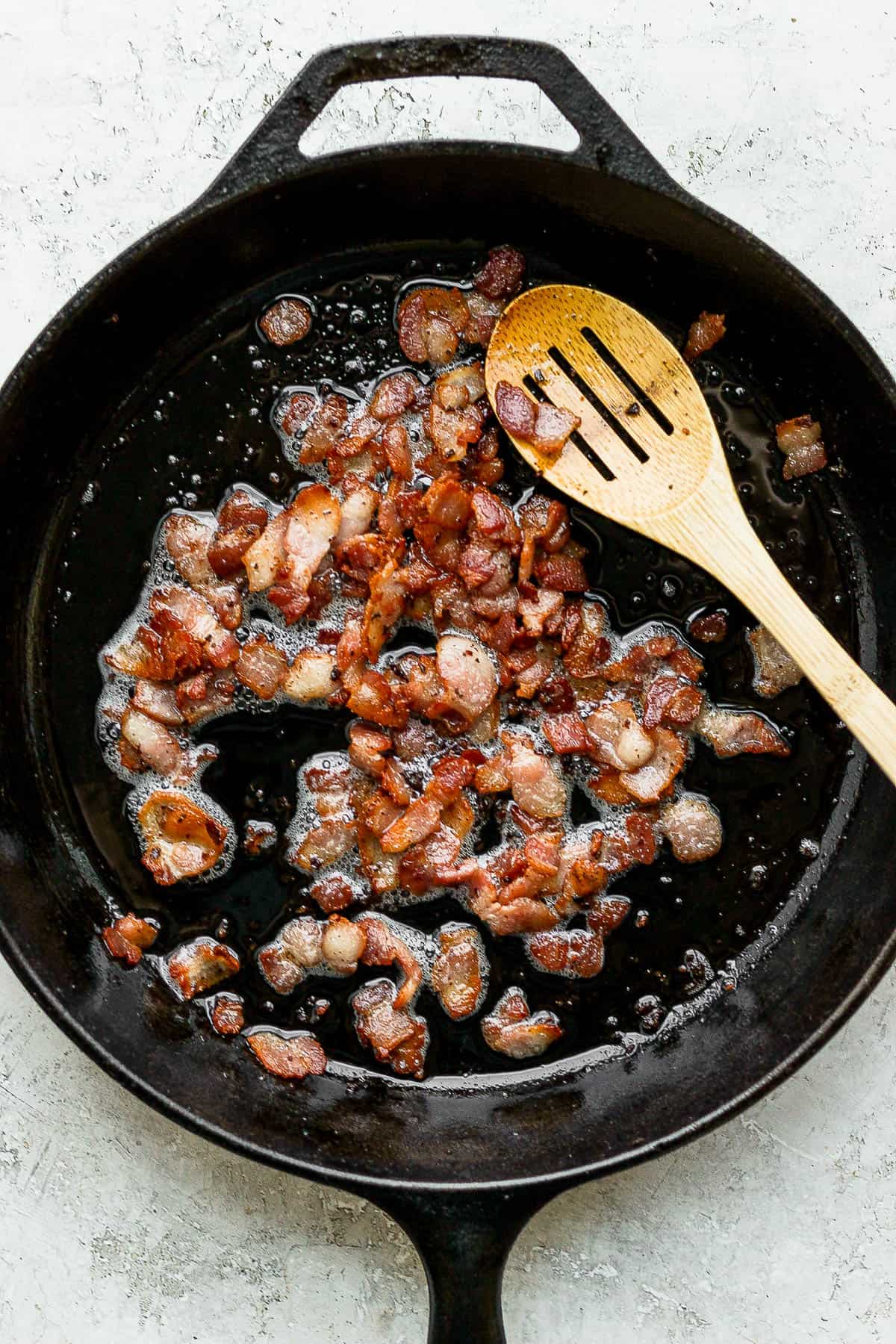 Bacon being cooked in a cast iron skillet with a wooden slotted spoon. 