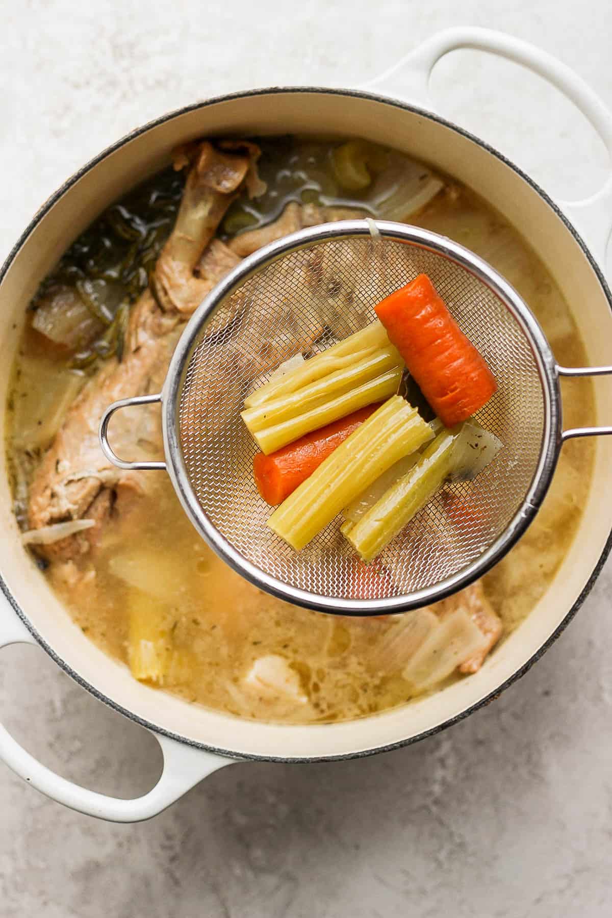 A small strainer over the top of the dutch oven with some celery and carrot inside showing how to strain the turkey stock. 