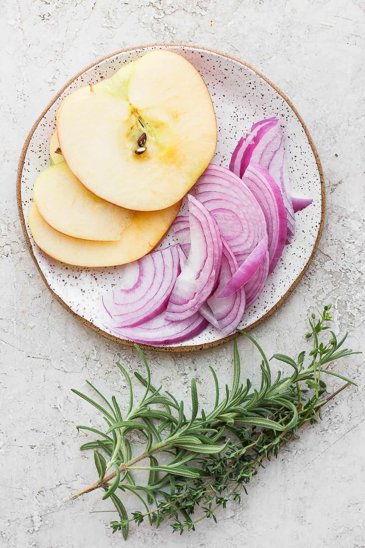 Sliced apple and red onion on a plate with sprigs of rosemary and thyme on the side.