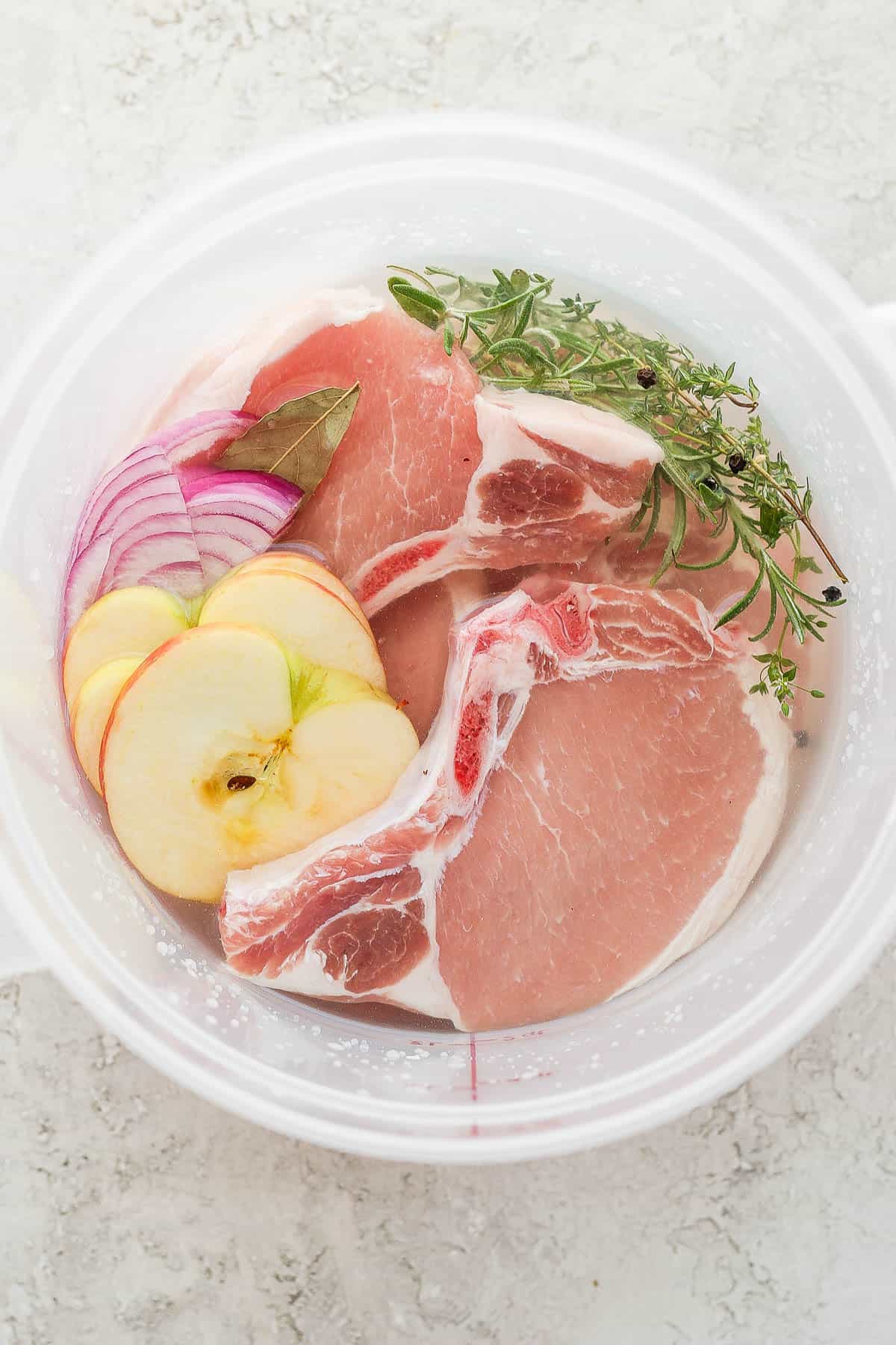 Brine mixture of water, salt, whole peppercorns, rosemary, thyme, bay leaf, onion, apple, and pork chops in a food safe container.