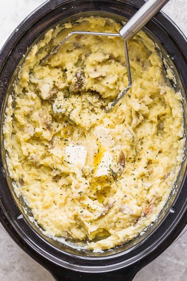 A large slow cooker filled with mashed potatoes and a potato masher sticking out.