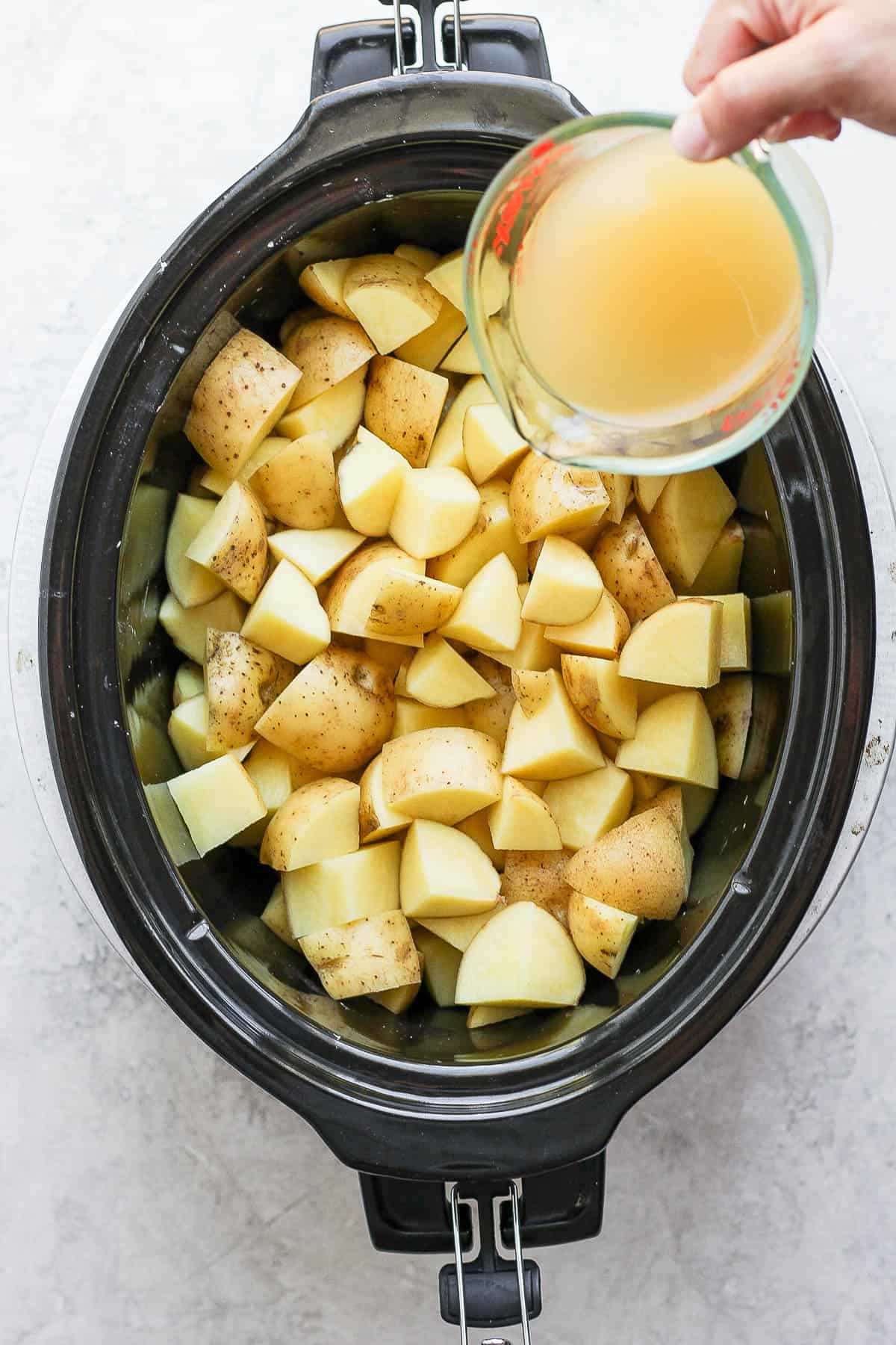 A measuring cup of chicken broth being poured over the top of the cubed yukon potatoes in the slow cooker.