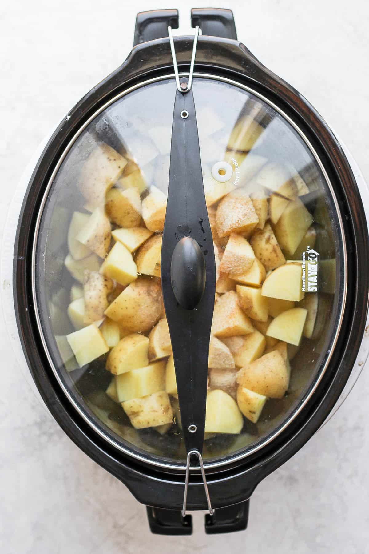 A slow cooker lid placed over the slow cooker containing the potatoes, broth, and seasonings. 
