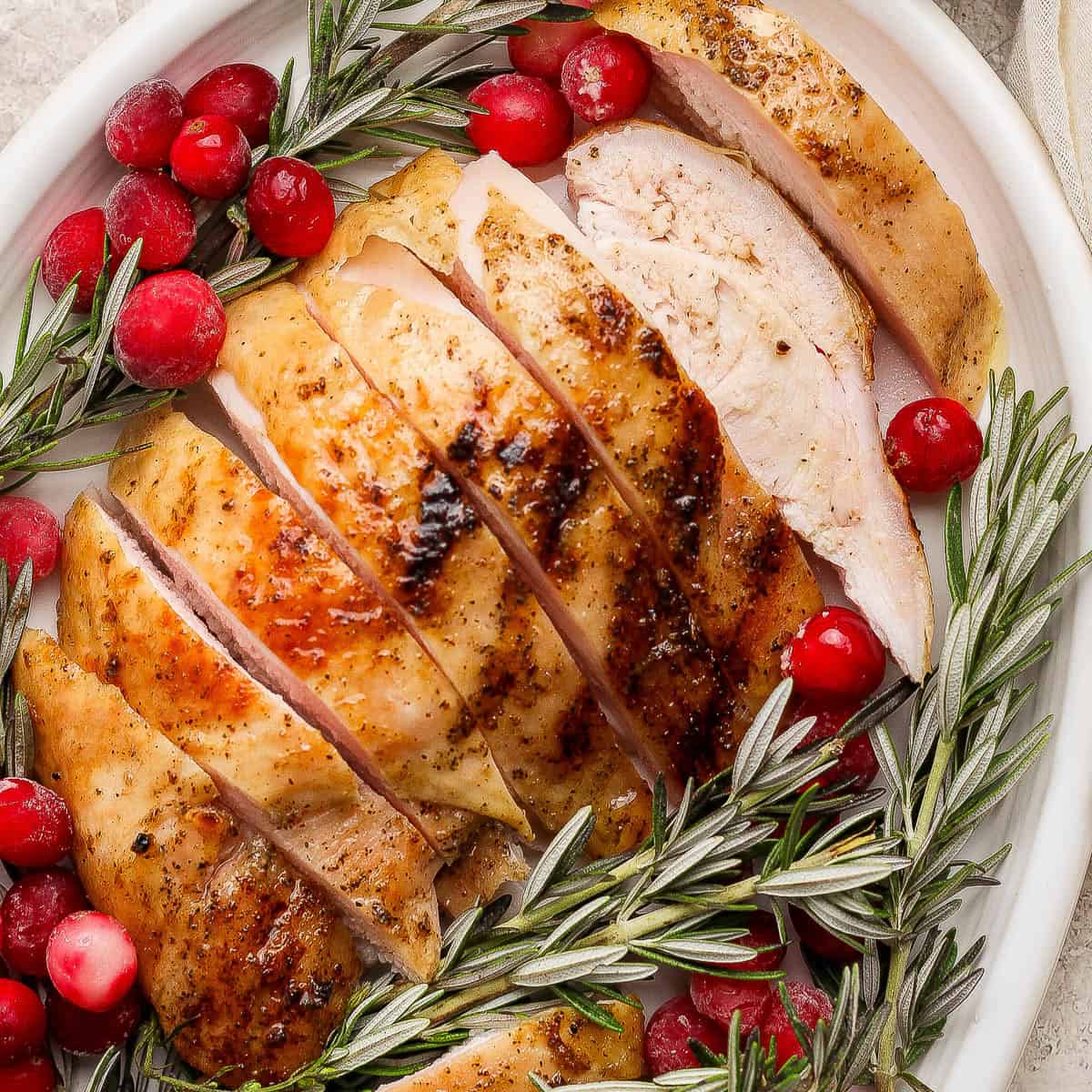 A platter with sliced smoked turkey breast on it along with cranberries and fresh rosemary.