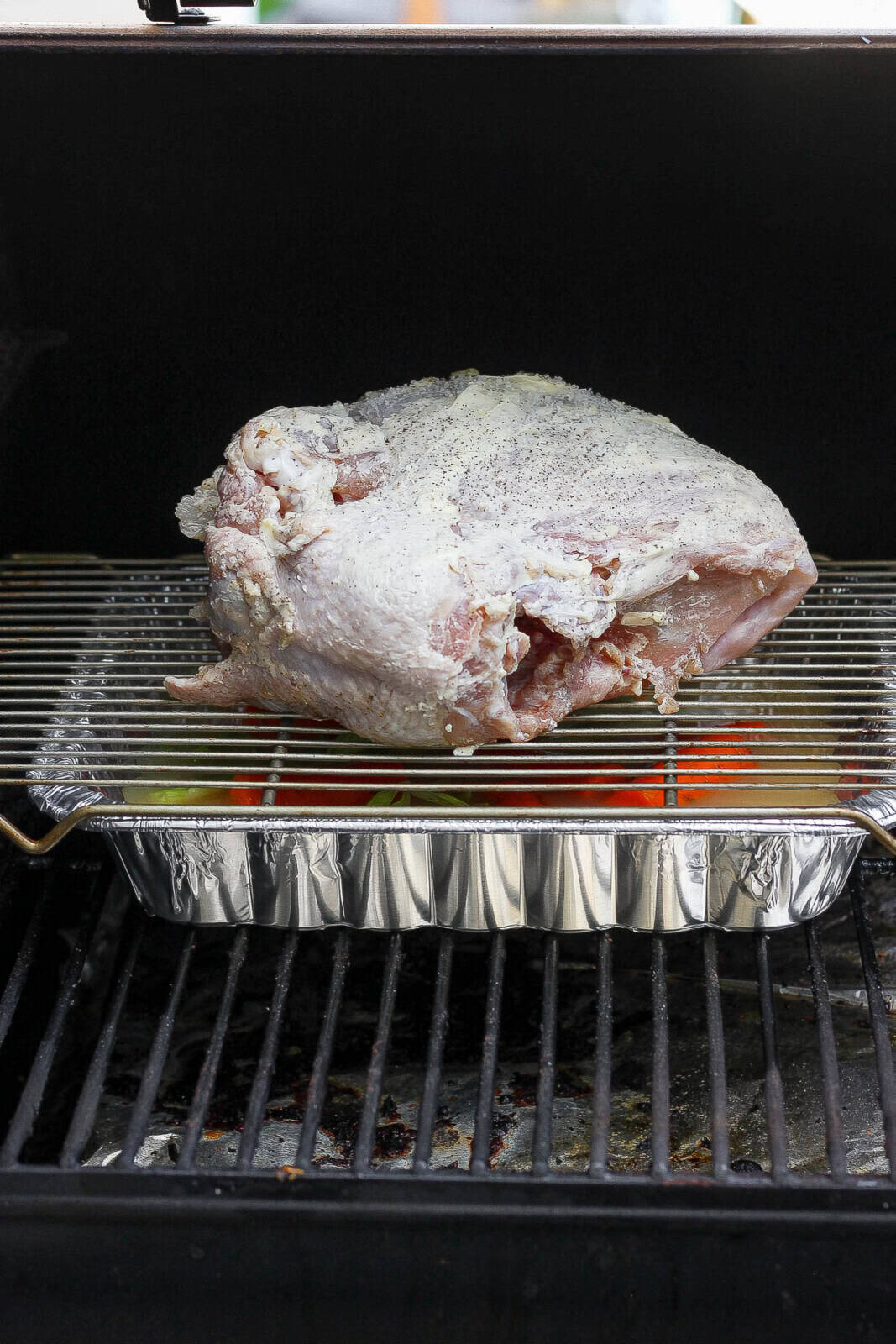 The prepared turkey breast on the smoker where it is on top of an aluminum pan to catch drippings with a wire rack in between to hold the turkey.