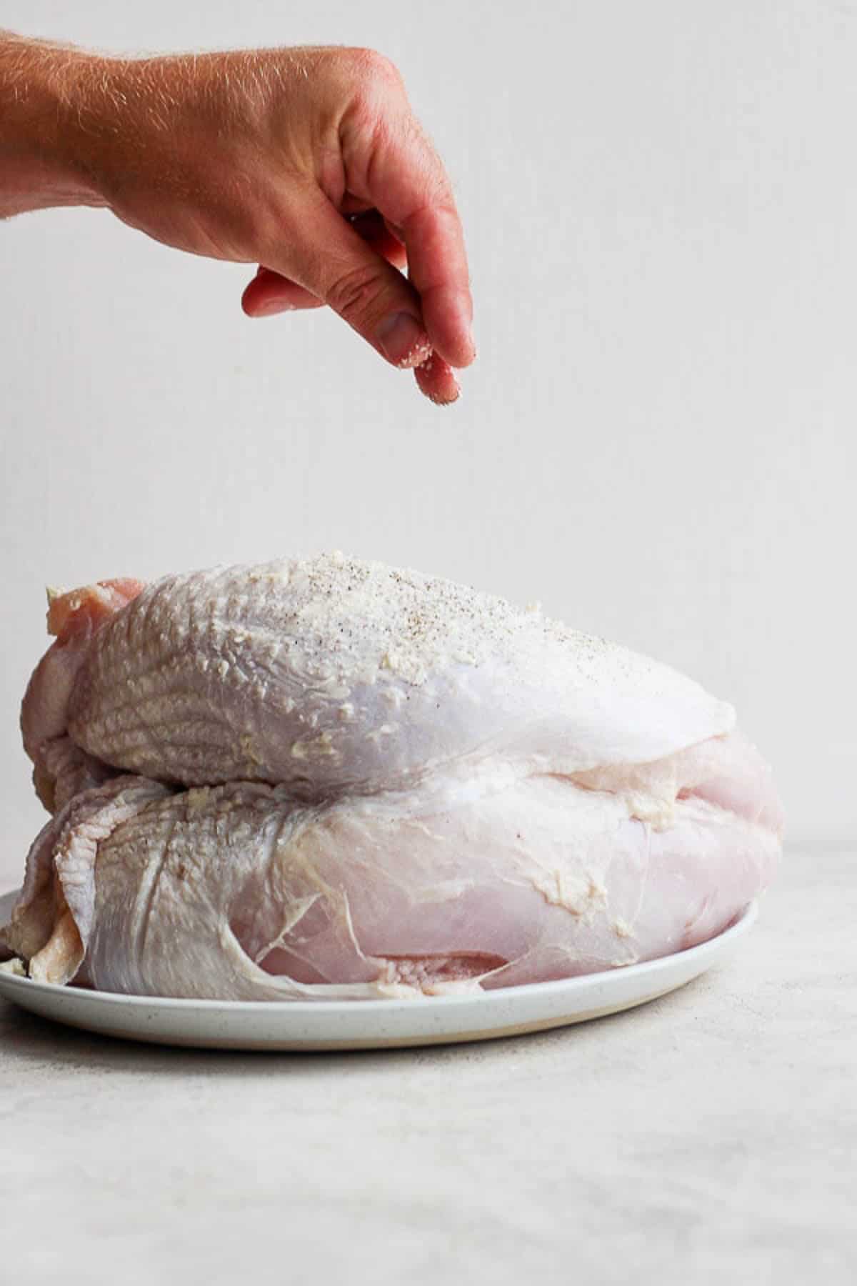 Someone sprinkling seasoning on a raw turkey breast.