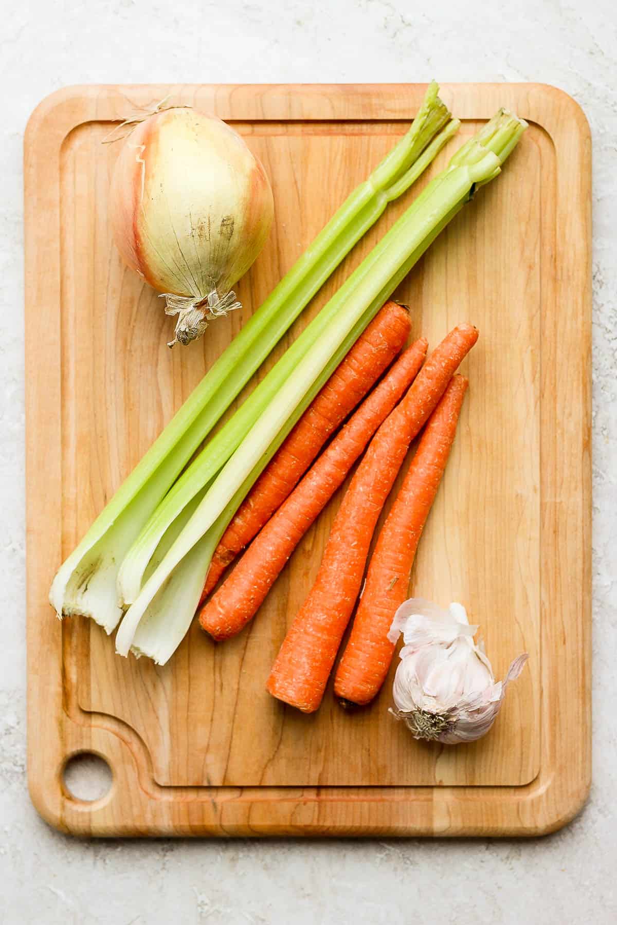 An onion, celery, carrots, and garlic on a cutting board.