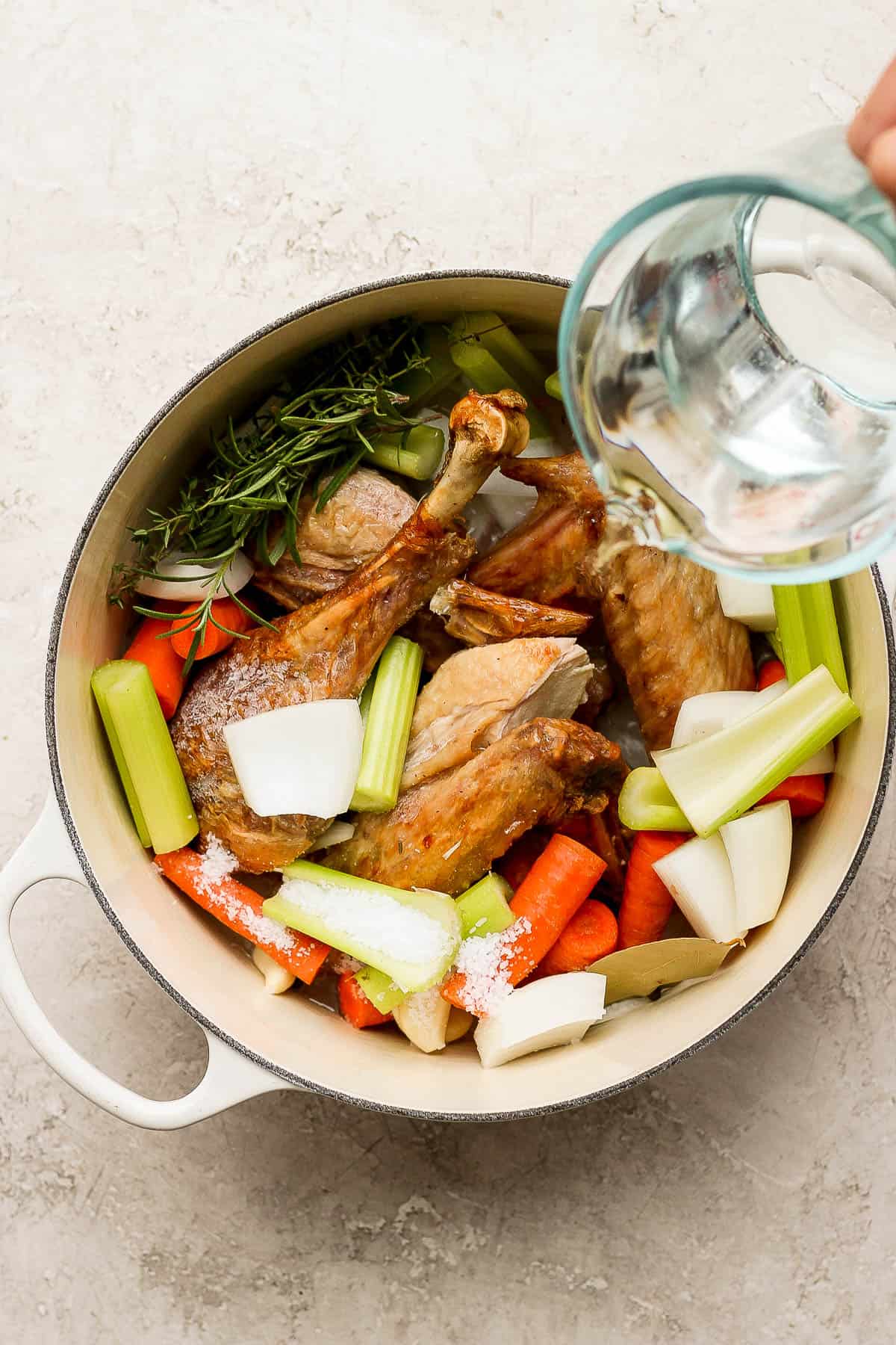 All meat, bones, vegetables, and herbs in a dutch oven with water being poured in.