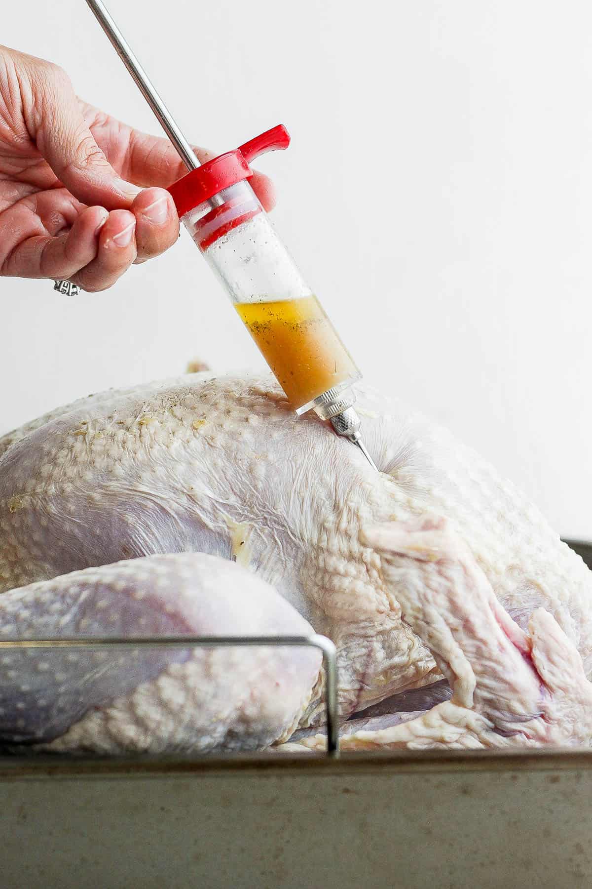 A raw turkey in a roasting pan being injected with a melted butter and herb mixture.