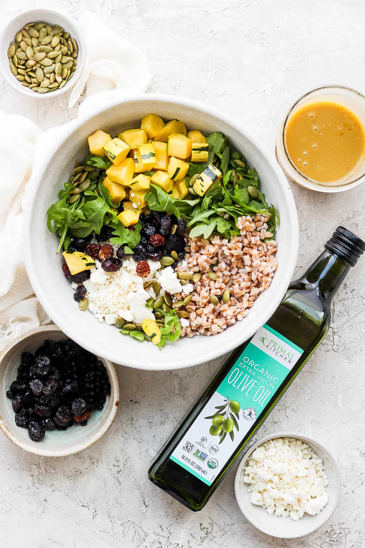 The farro salad in a large mixing bowl with the jar of dressing, an olive oil bottle, pepitas, & dried cranberries on the side.