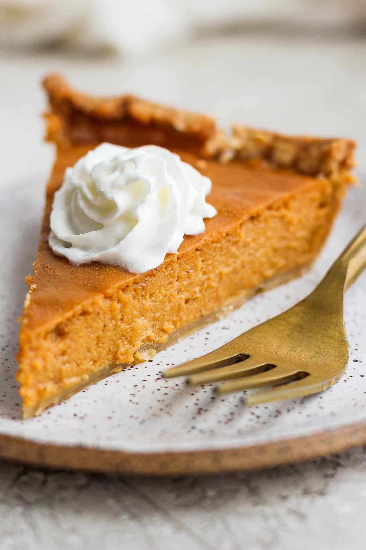 A close up of a piece of pumpkin pie with a dollop of whip cream on a plate alongside a golden fork.