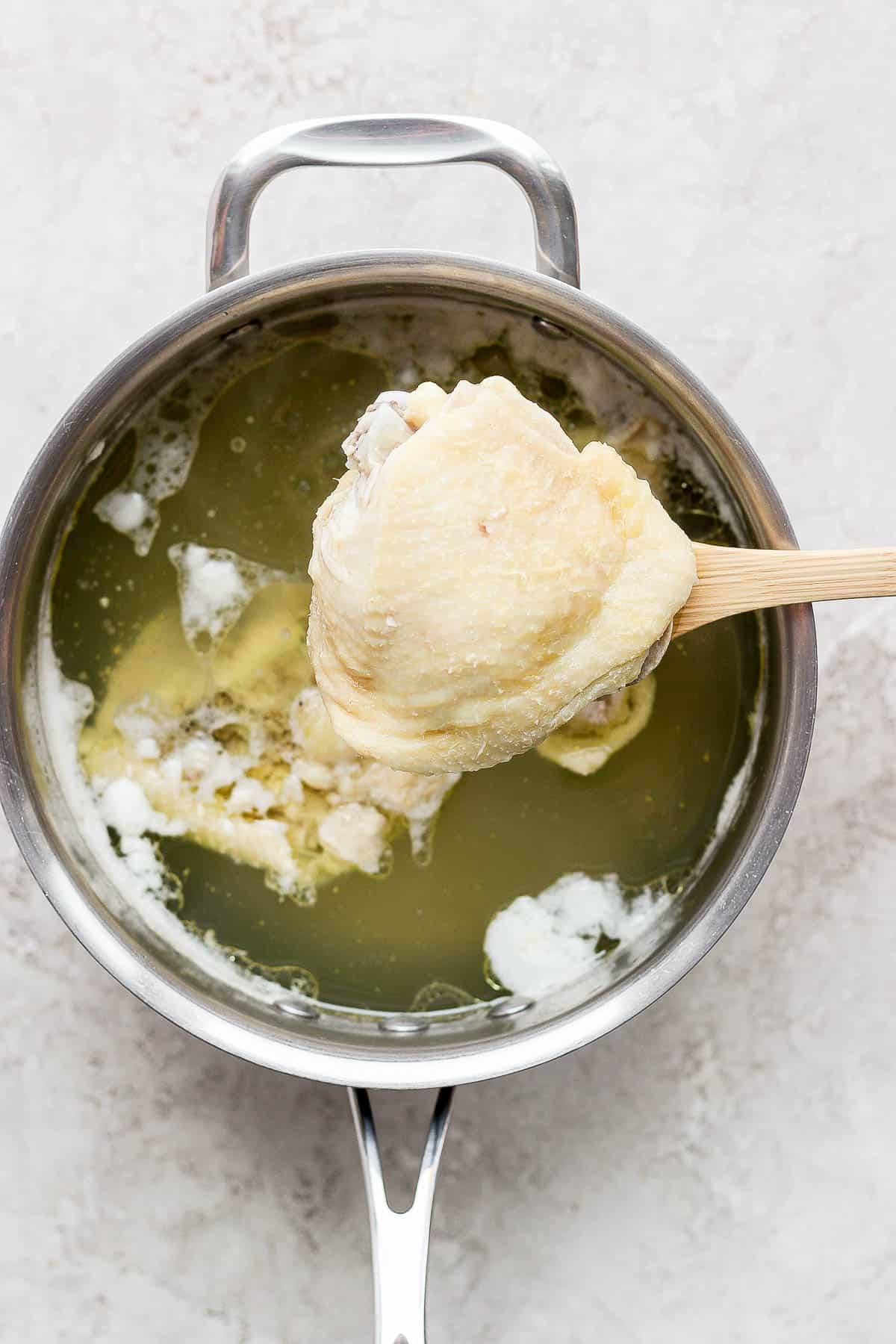 A spoon scooping out a boiled chicken thigh out of a pot of water.