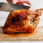 A cooked bone-in turkey breast on a cutting board with someone just about to carve it with a chef's knife.