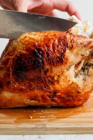 A cooked bone-in turkey breast on a cutting board with someone just about to carve it with a chef's knife.