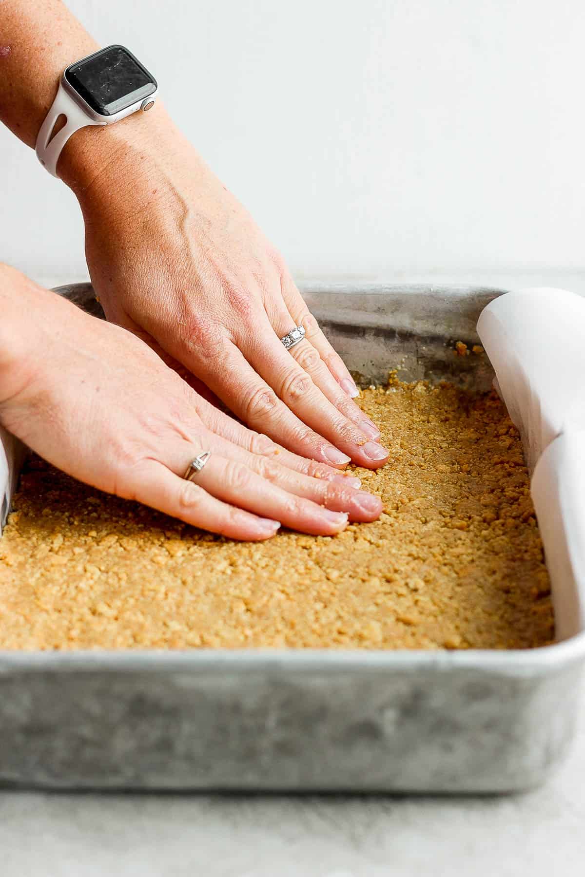 Hand pushing the graham cracker crust into a parchment-lined baking pan.