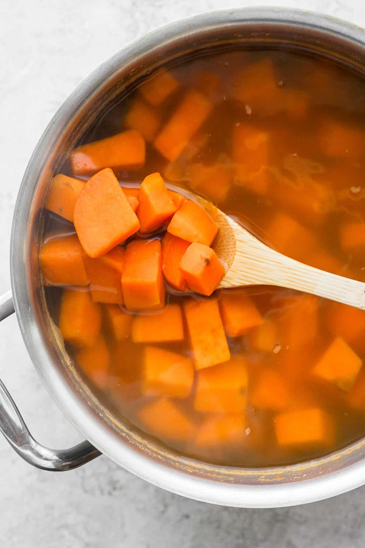 Cubed and peeled sweet potatoes boiled in a large pot.  A wooden spoon lifts a few out to show that they've been cooked.