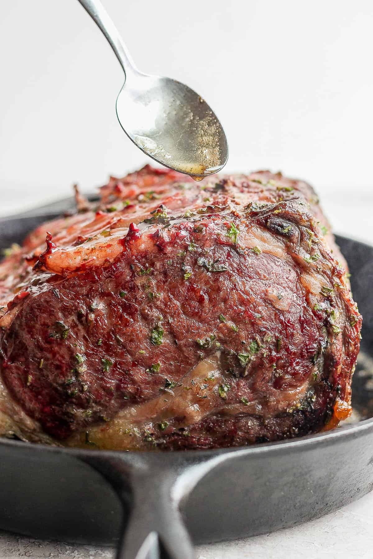 Prime rib being seared in a cast iron skillet and a spoon scooping the rendered fat over the top.
