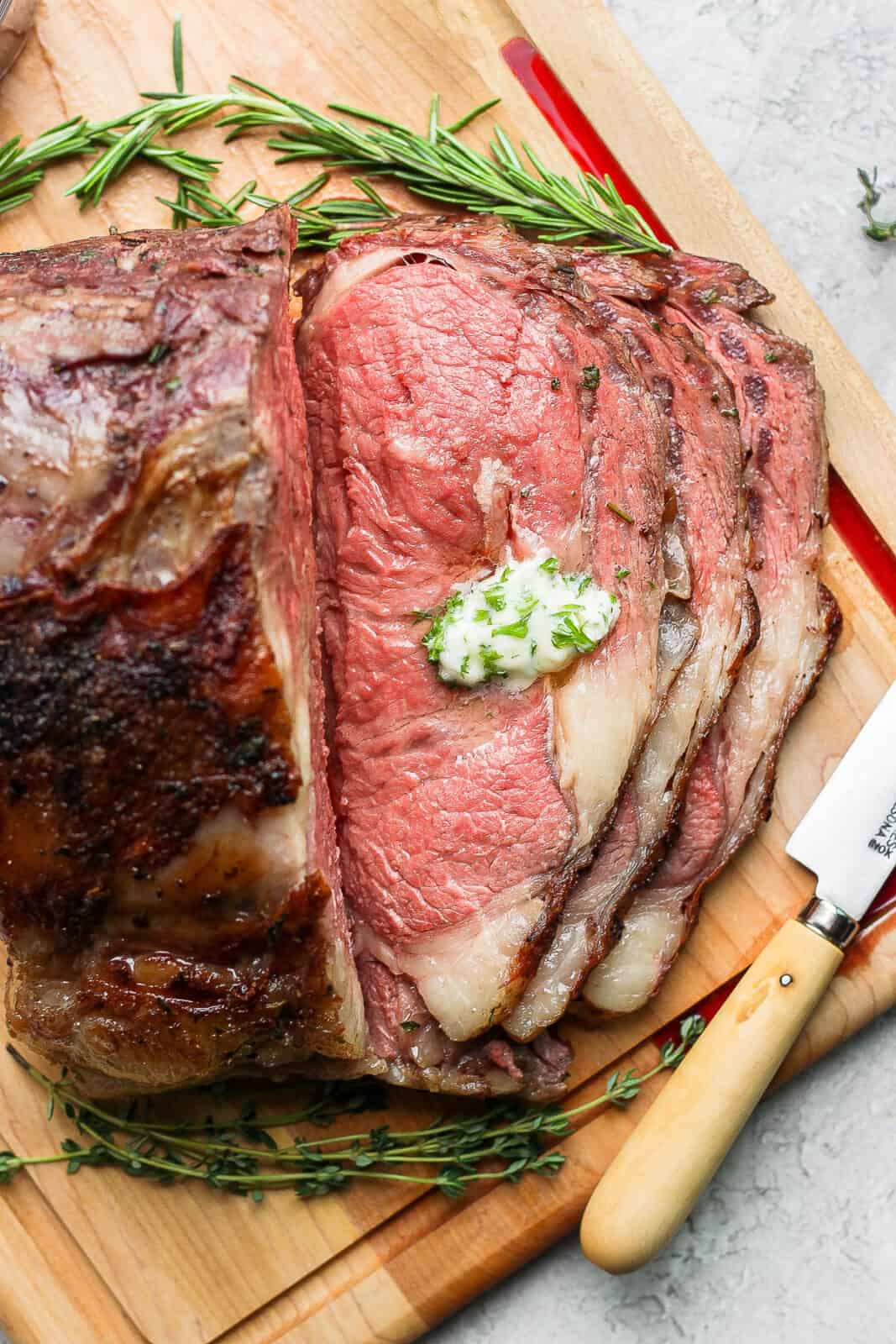 A medium rare smoked prime rib on a cutting board with 3 slices cut into it and some herbed butter on the top slice.