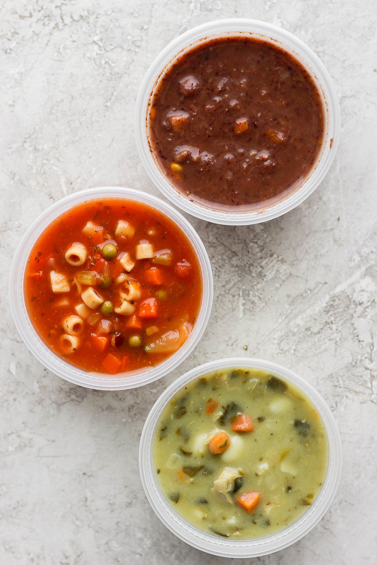 Three soup containers with the lids removed showing three different varieties of soup: a black bean soup, a minestrone soup, and a chicken gnocchi soup.