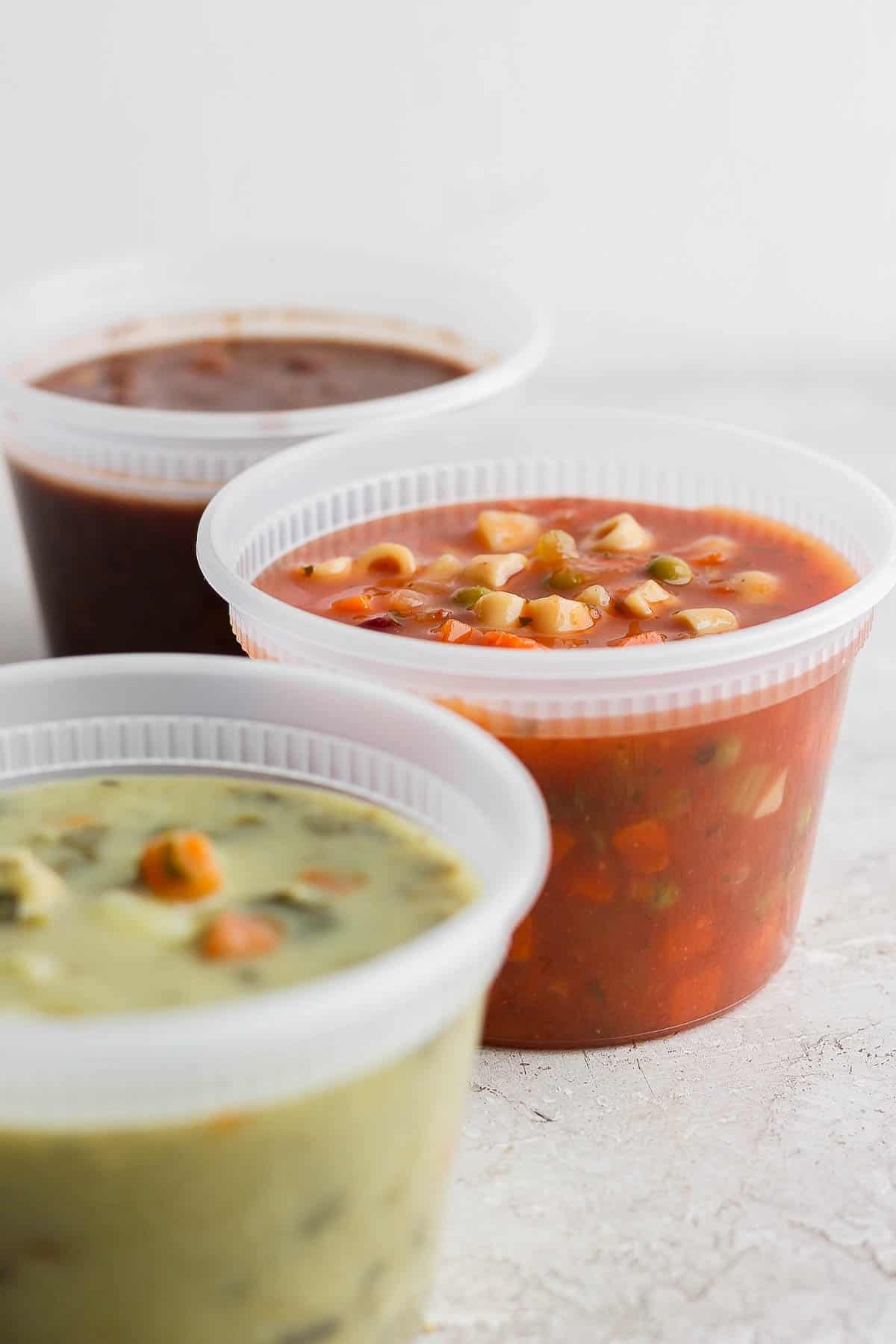 Three soup containers with the lids removed showing three different varieties of soup: a black bean soup, a minestrone soup, and a chicken gnocchi soup.
