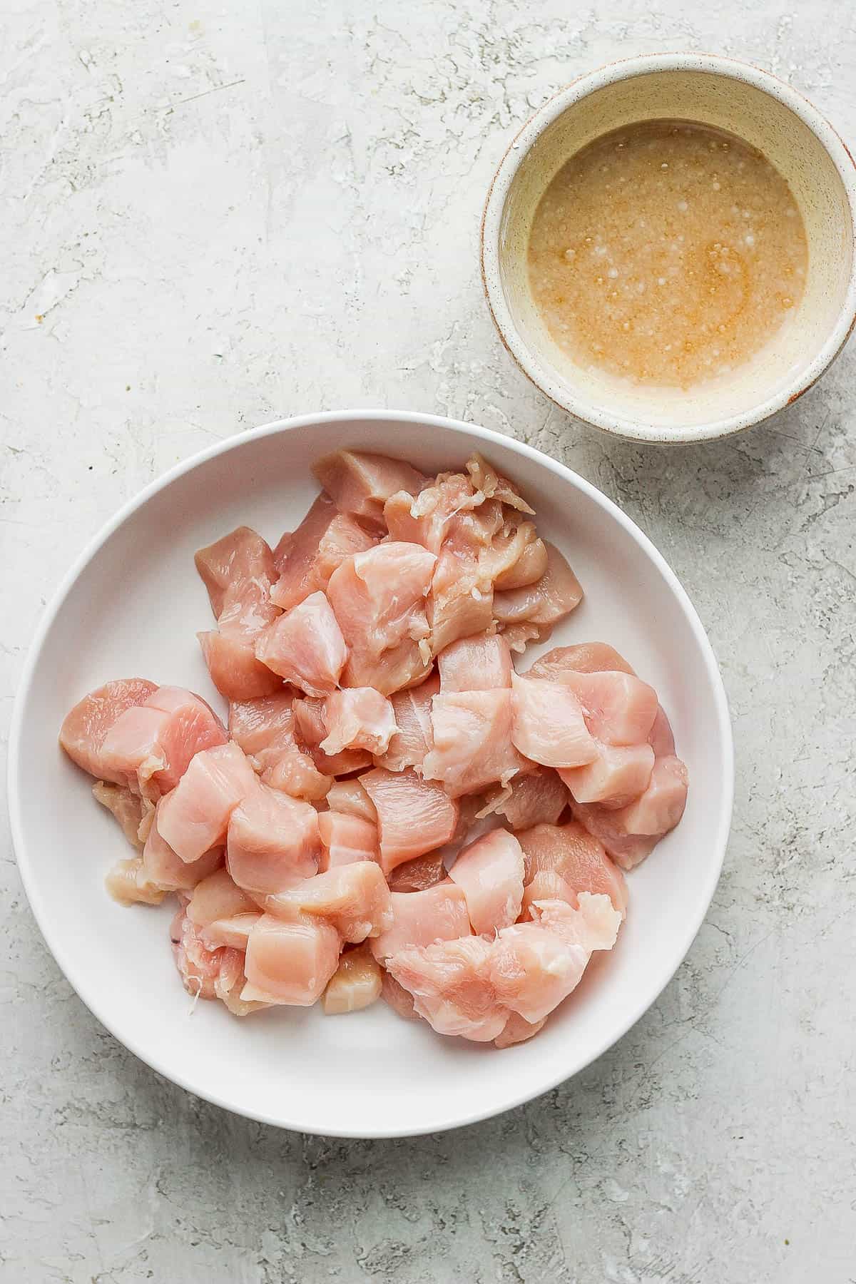 Raw chicken chunks in a white bowl and the marinade in another small bowl.
