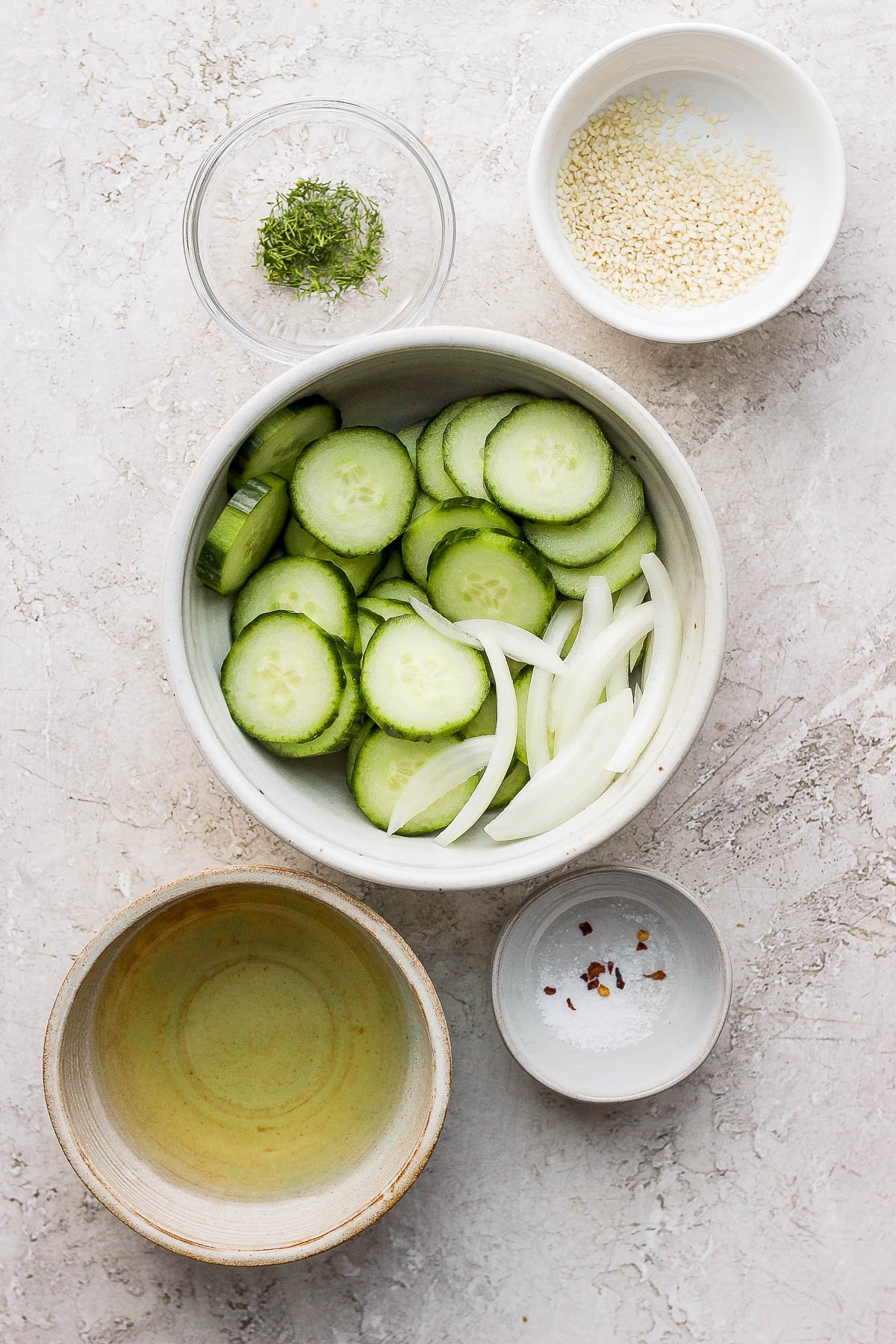 Five separate bowls all containing measured and prepped ingredients: fresh dill, sesame seeds, sliced onion and cucumber, apple cider vinegar and rice vinegar, and finally salt and red pepper flakes.