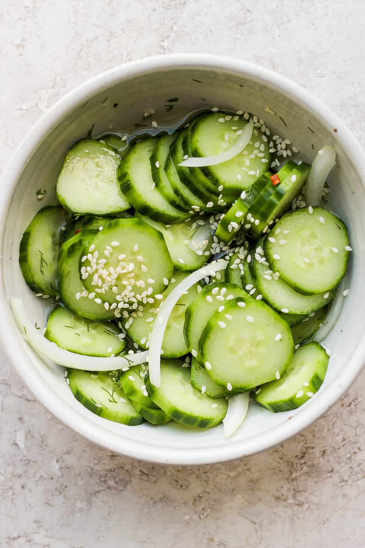 Marinated cucumbers in a bowl with onions, sesame seeds, dill, and red pepper flakes. 