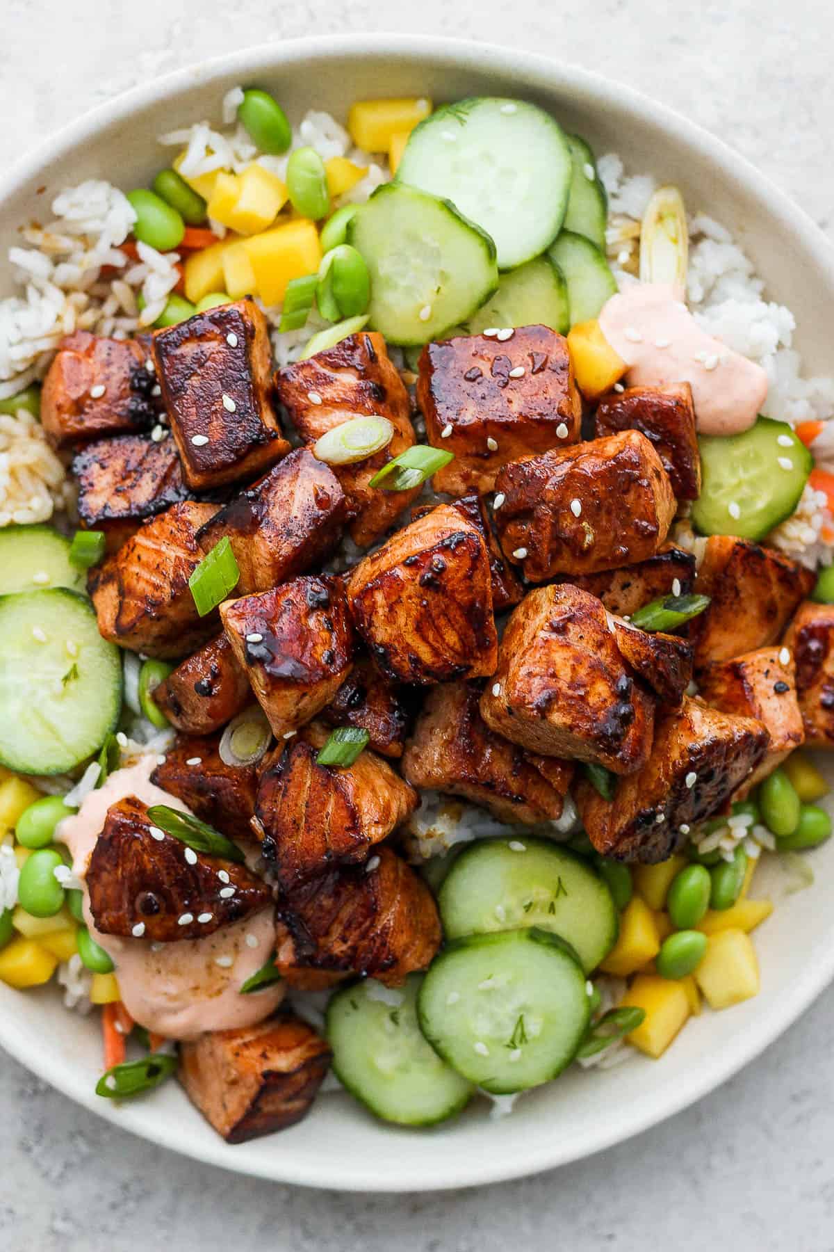 A bowl of rice topped with chopped mango, edamame, marinated cucumbers, green onions, salmon, and sriracha mayo.