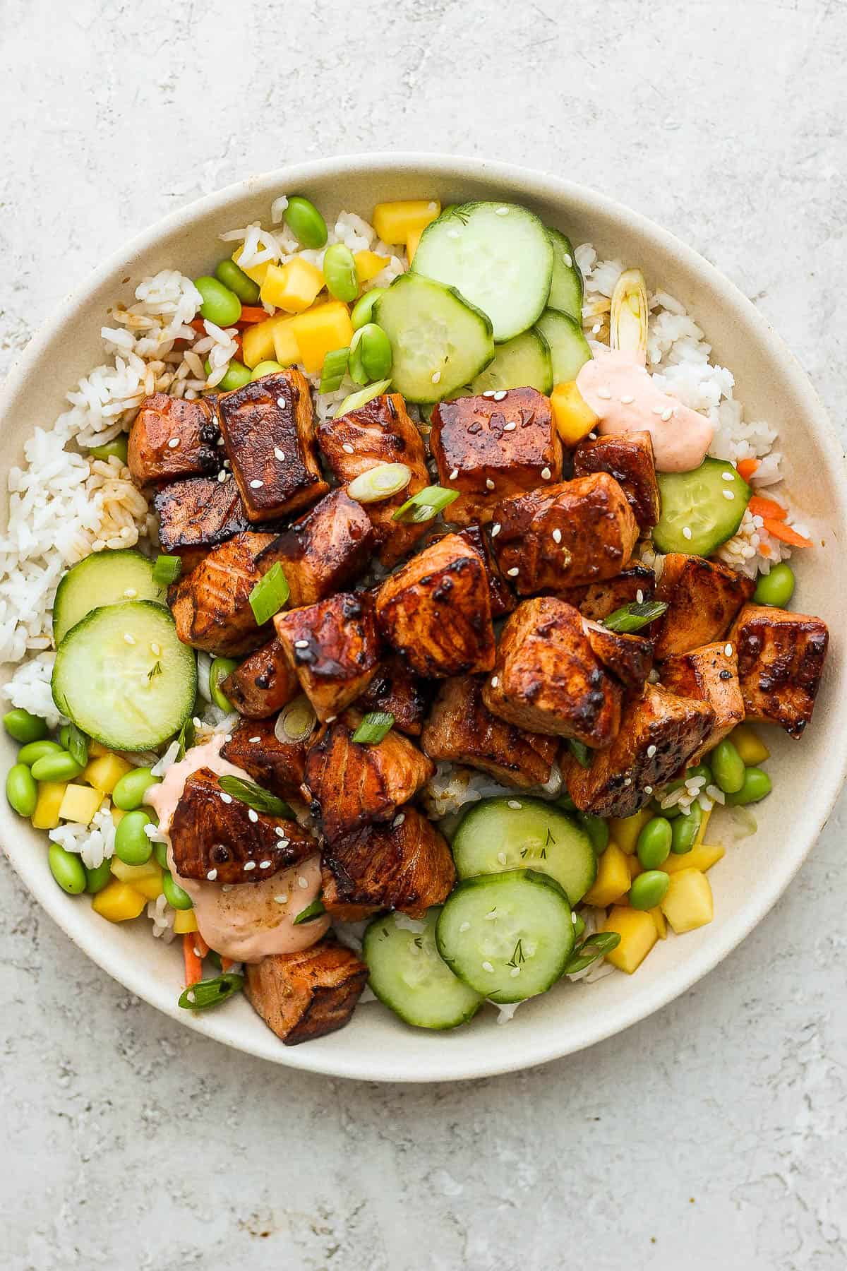 A bowl of rice topped with chopped mango, edamame, marinated cucumbers, green onions, salmon, and sriracha mayo.