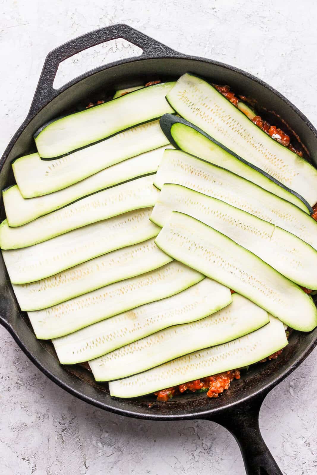 A layer of zucchini slices in a cast iron skillet to make zucchini lasagna with meat sauce.