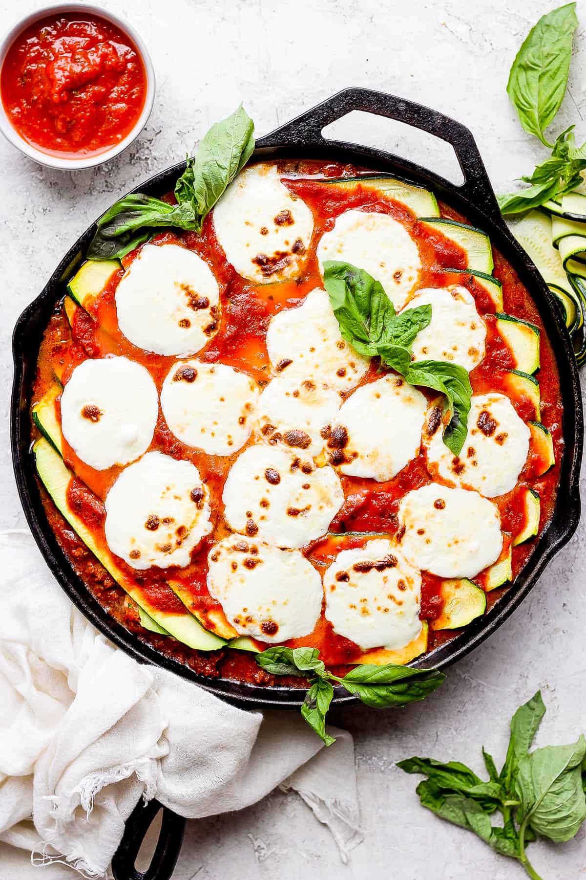 A zucchini lasagna baked in a cast iron skillet garnished with fresh basil leaves.  A small bowl of marinara sauce is off to the side. 